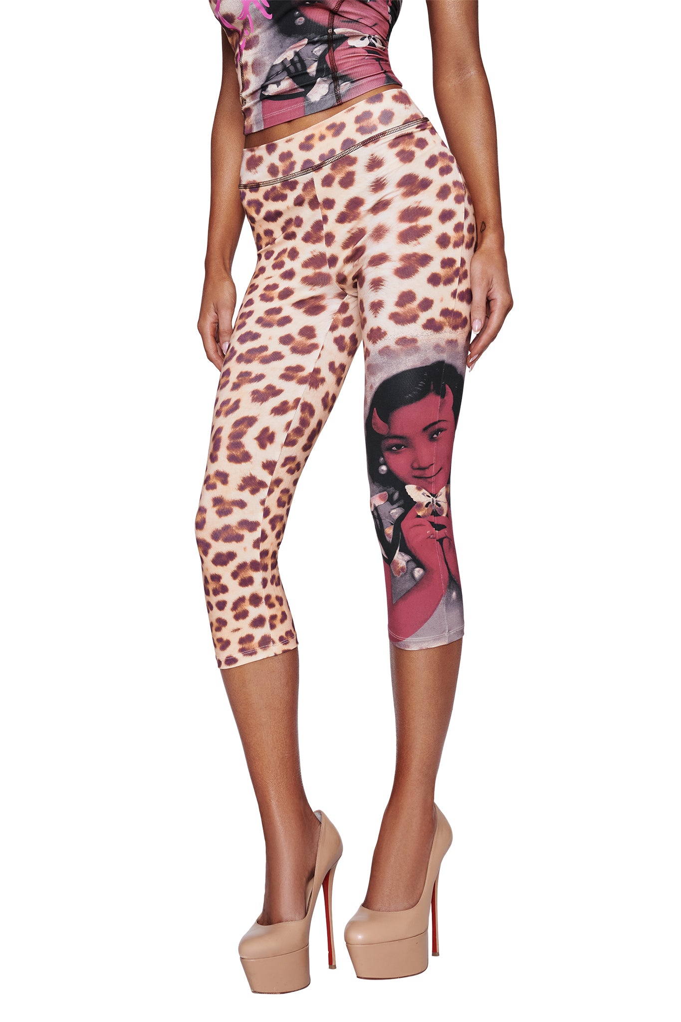 A woman wearing I.AM.GIA's T-REX BLARE CAPRI - WILD CARD capri pants featuring a cheetah print and cartoon woman illustration, paired with a patterned top and nude high heels, stands against a white background.