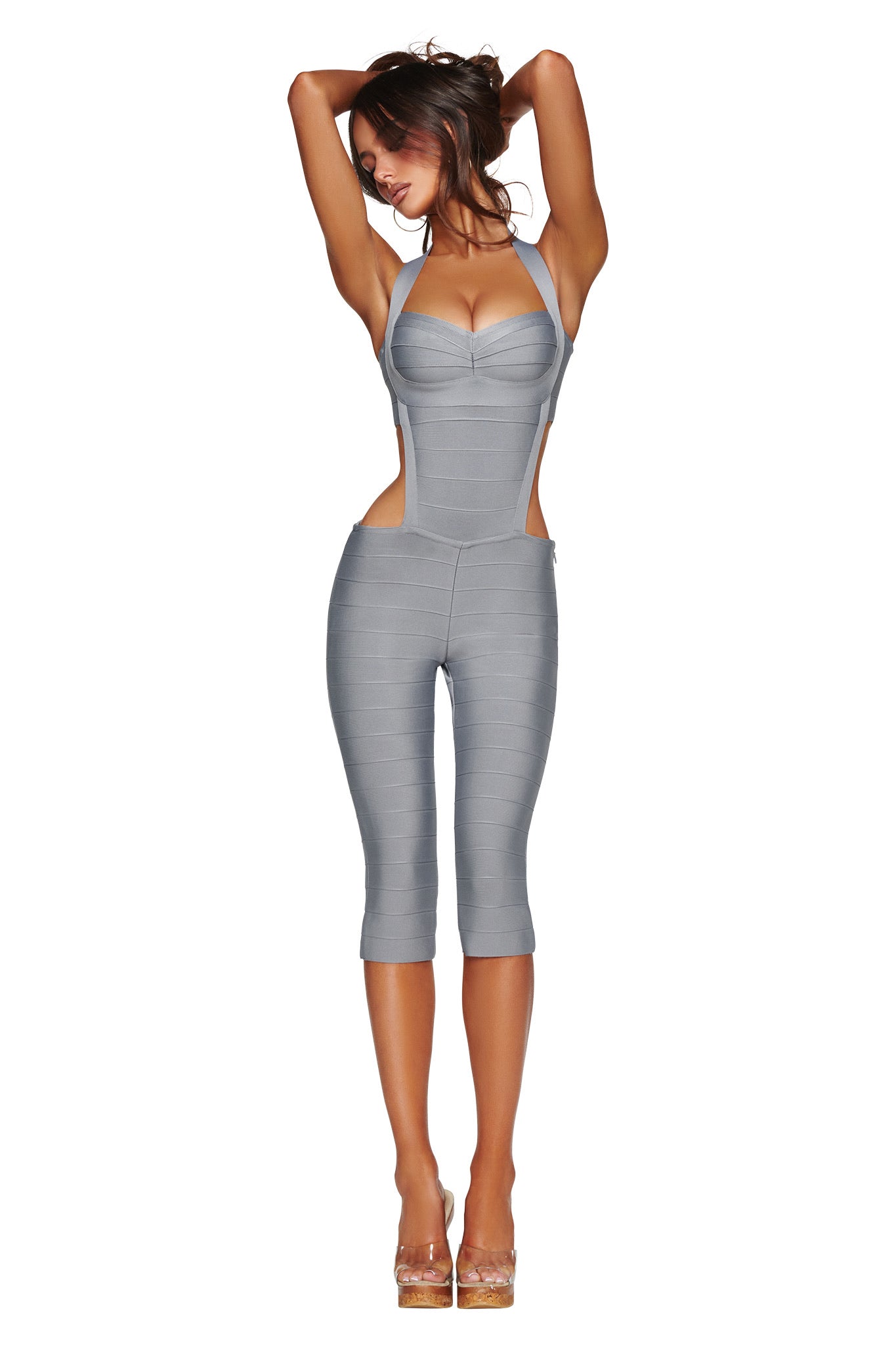 A woman stands with arms raised above her head against a white background, wearing the I.AM.GIA EVA BANDAGE CAPRI JUMPSUIT in grey—a fitted, halter neck style with cutout sides—paired with clear high heels.