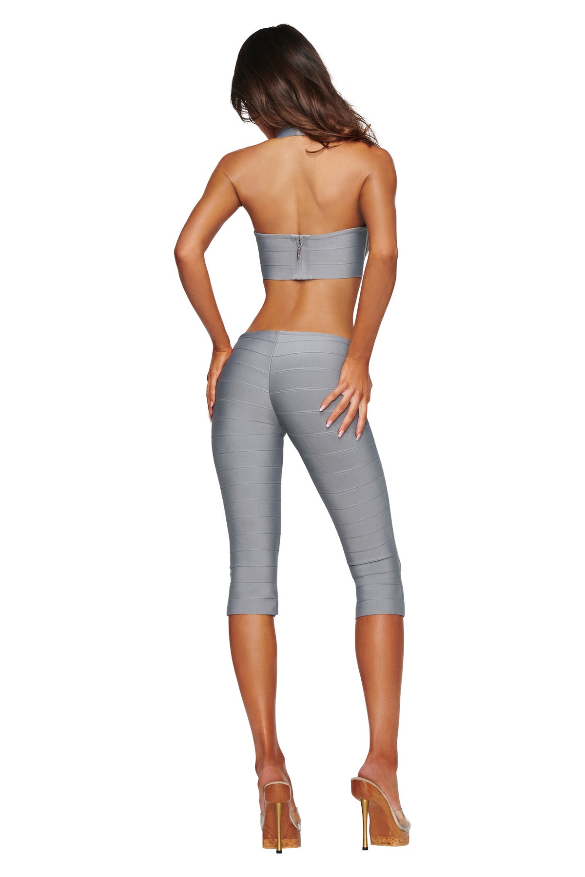 A woman with long hair stands facing away against a white background, wearing the I.AM.GIA EVA BANDAGE CAPRI JUMPSUIT in grey—a strapless, bodycon design featuring a crop top and matching high-waisted capri pants—paired with high-heeled clear shoes.