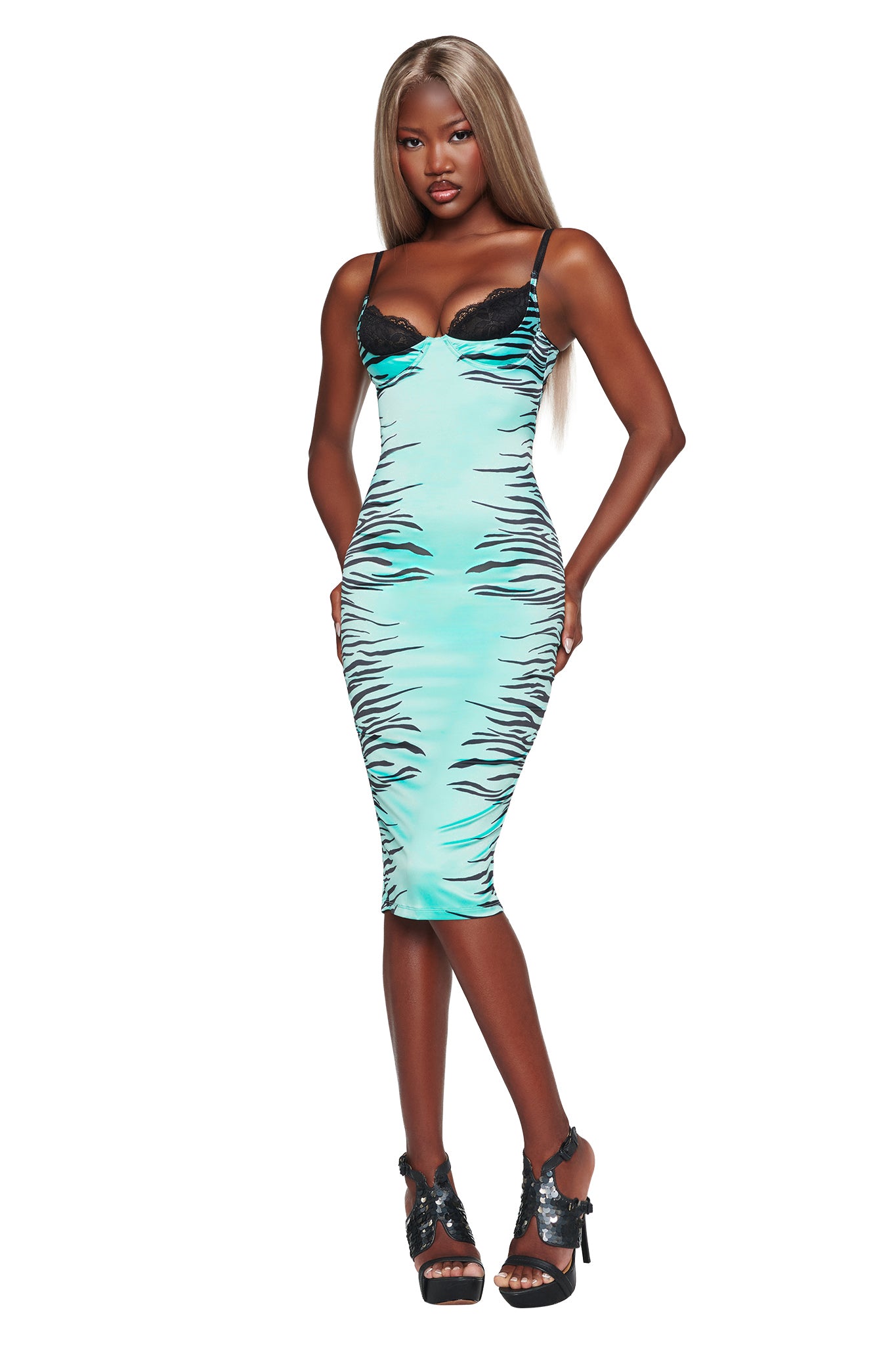 A woman stands against a white background with her hands on her hips, dressed in the I.AM.GIA CASSY MIDI DRESS in Aqua Fine Line Zebra—a fitted, sleeveless dress featuring an aqua zebra print with black tiger stripes and black lace trim—paired with black high-heeled sandals.