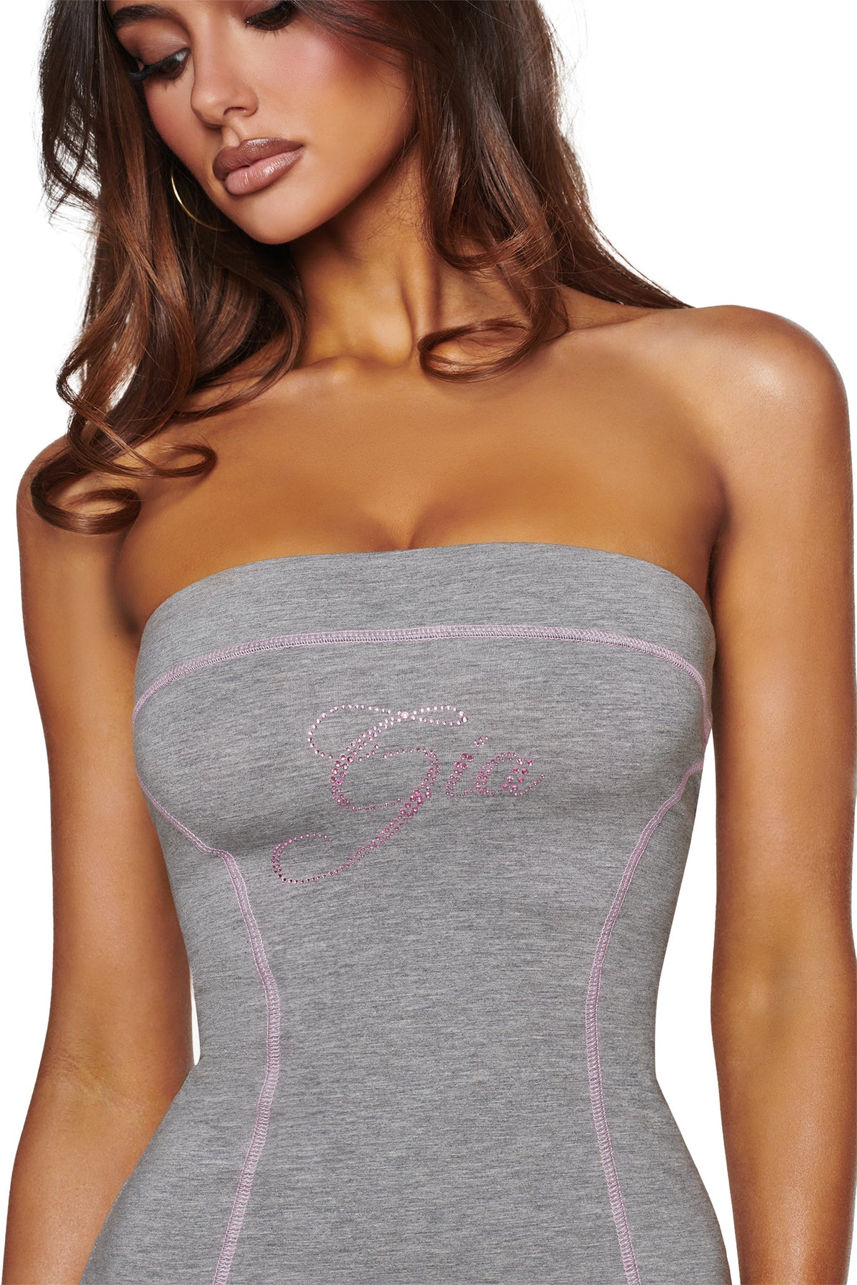 A woman is wearing the I.AM.GIA BLARE TUBE MINI DRESS in heather grey, a strapless and fitted piece featuring pink stitching and the word “Gia” spelled out in pink rhinestones across the chest. Her face is partially visible, and she has wavy brown hair.