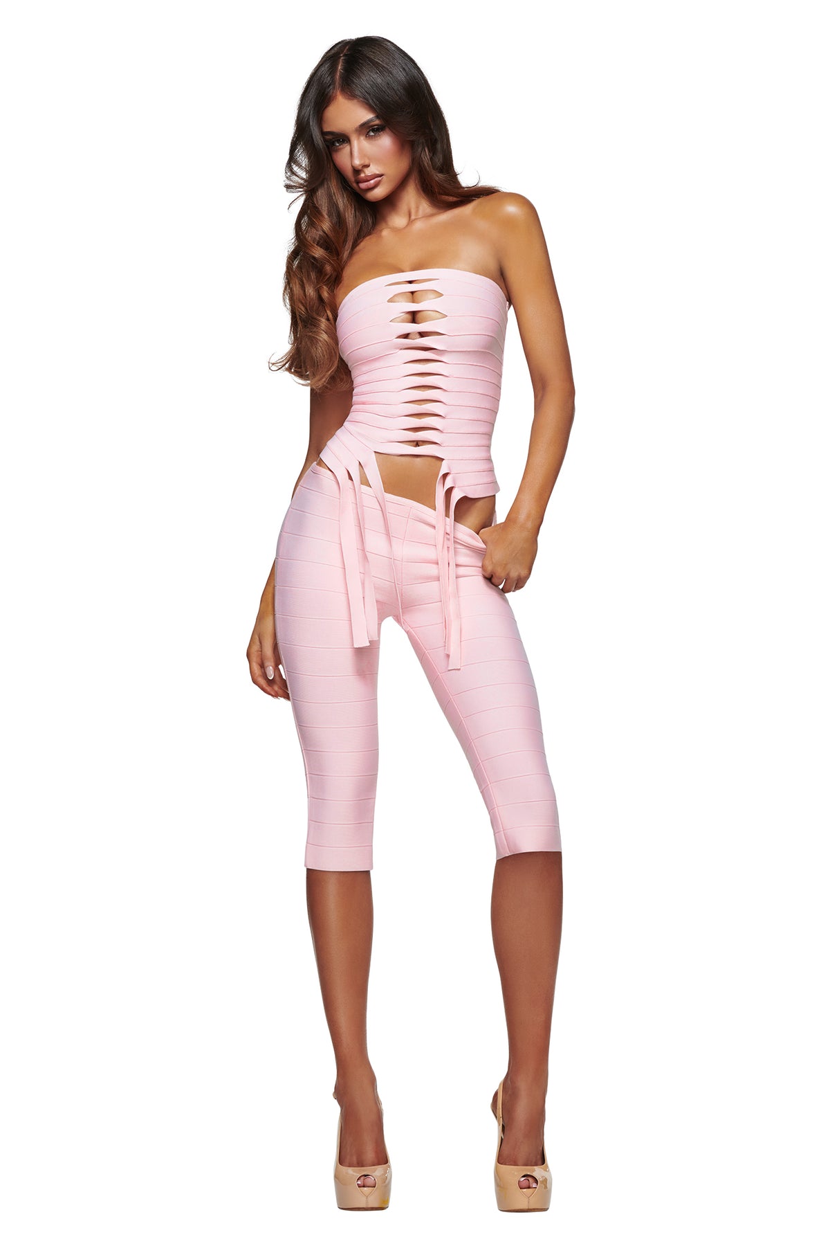 Model wears the I.AM.GIA MAEVE BANDAGE CAPRI in baby pink, featuring a bodycon fit with a matching light pink, strapless, cut-out top, styled with high heels and posed against a white background.