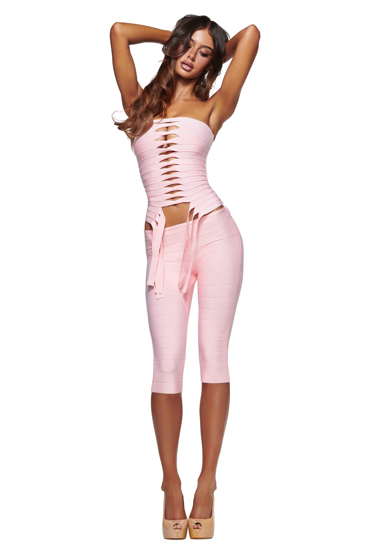 A woman poses against a white background, arms raised behind her head, wearing the I.AM.GIA MAEVE BANDAGE CAPRI in baby pink—a sleek bodycon set featuring a light pink, strapless cut-out top and matching capri pants—styled with high heels.
