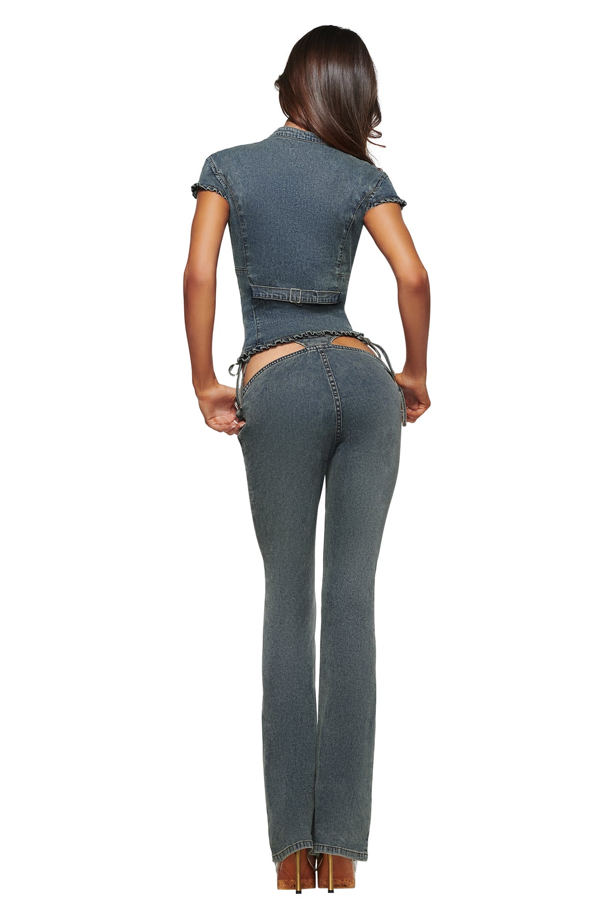 A woman stands with her back to the camera, wearing the I.AM.GIA JET DENIM TOP in Faded Indigo, paired with distinctive indigo denim pants featuring exposed cutouts at the hips—all set against a white background for a striking nod to bold Y2K style.