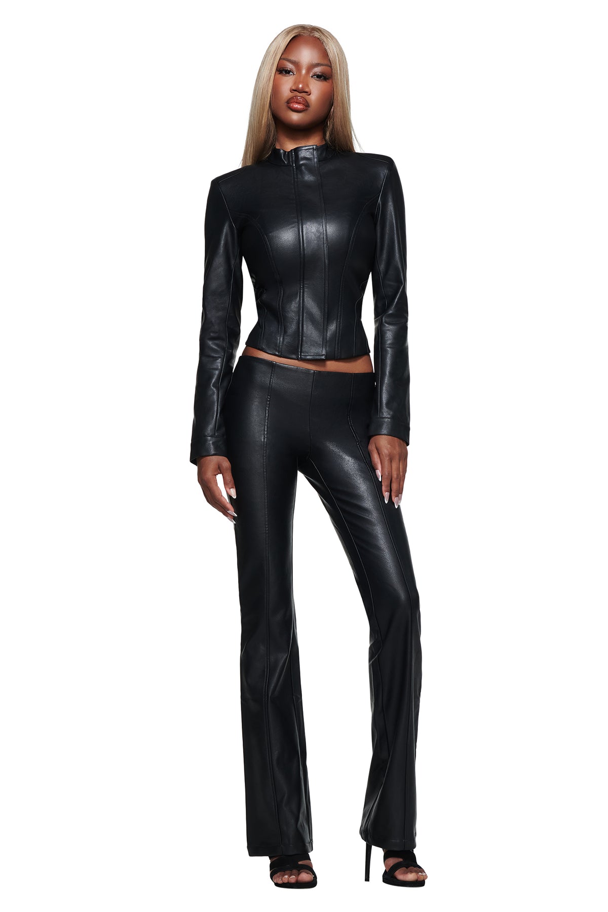 A woman stands confidently against a white background wearing the I.AM.GIA DUA PU JACKET in black, a fitted faux leather piece featuring a chic faux fur collar, styled with matching black leather pants and high-heeled sandals.