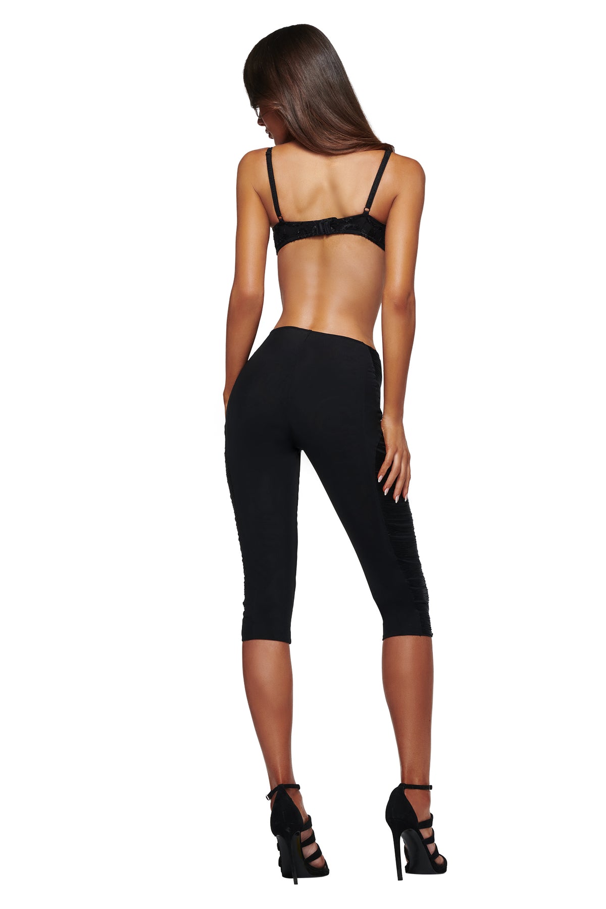 A woman with long brown hair stands facing away against a white background, wearing the I.AM.GIA VERON BRA in black, paired with black capri pants and black high heels.
