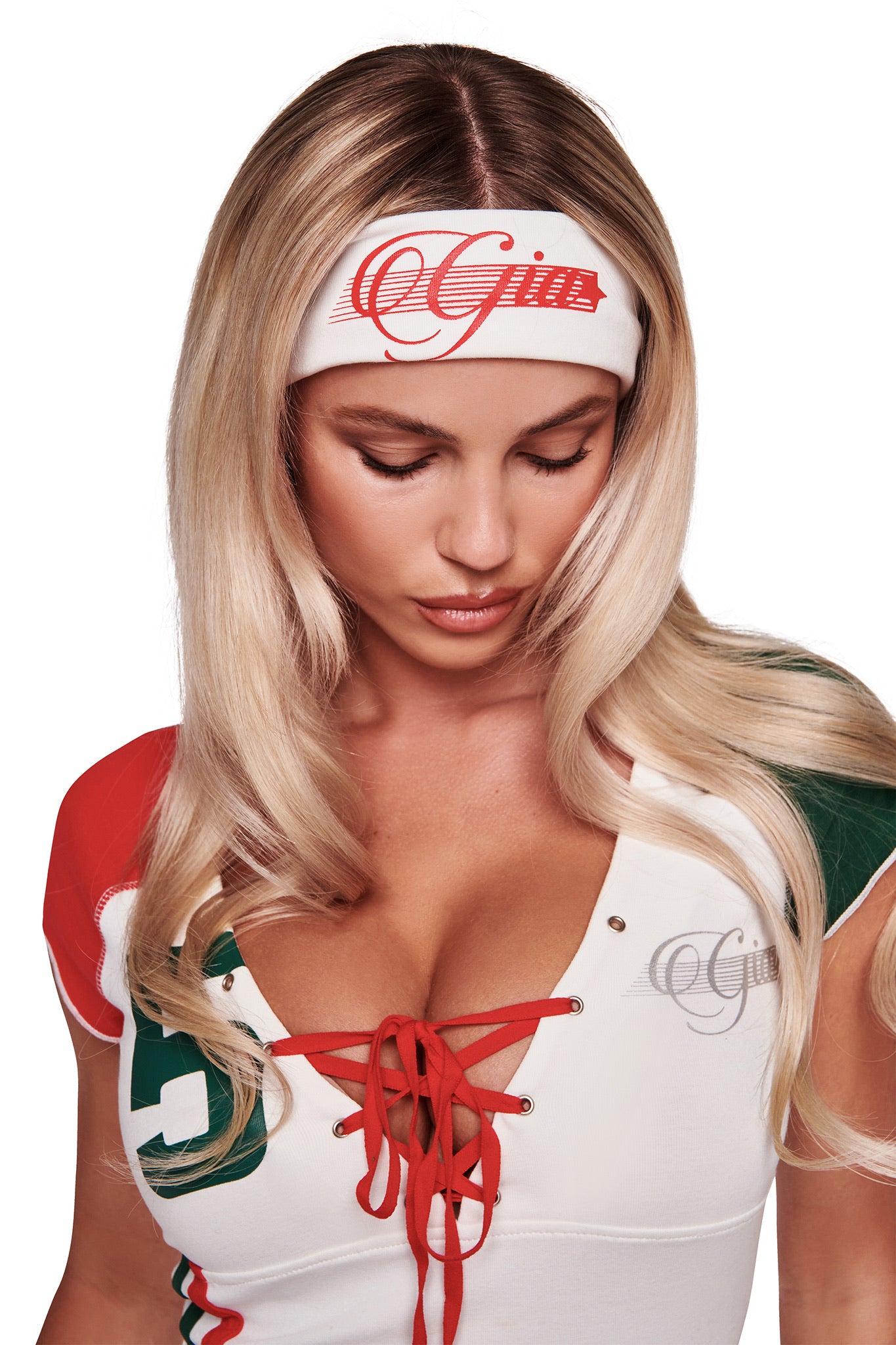 A woman with long blonde hair looks down, wearing the I.AM.GIA NOVIA SPORT HEADBAND in white. Her white top features red laces with green, red, and silver accents, set against a plain white background.