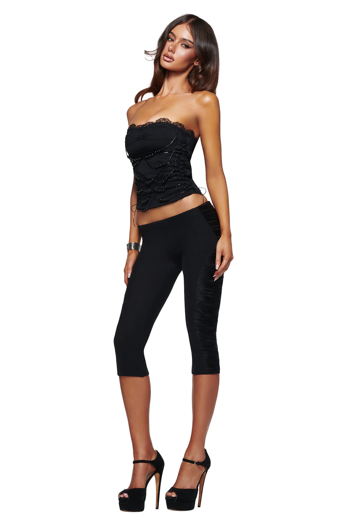 A woman poses against a white background, looking at the camera with one hand on her hip and showcasing her form-fitting silhouette in the I.AM.GIA DIXI CAPRI - BLACK leggings paired with a strapless black top and high heels.