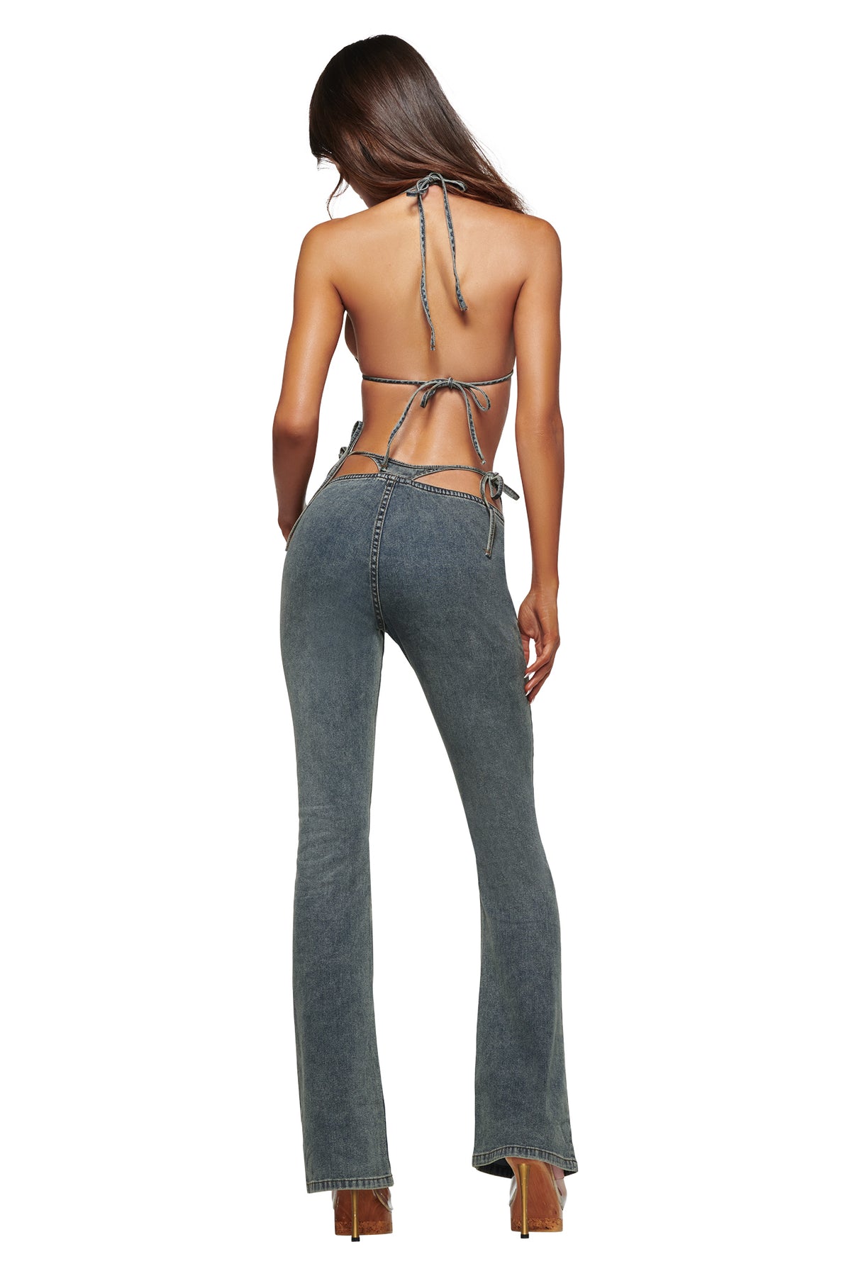 A woman faces away from the camera, wearing the I.AM.GIA JET G-STRING DENIM PANT in Faded Indigo—a pair of Y2K-inspired, high-waisted, flared jeans featuring side chains and a subtle G-string detail—styled with a backless halter top and high heels against a white background.