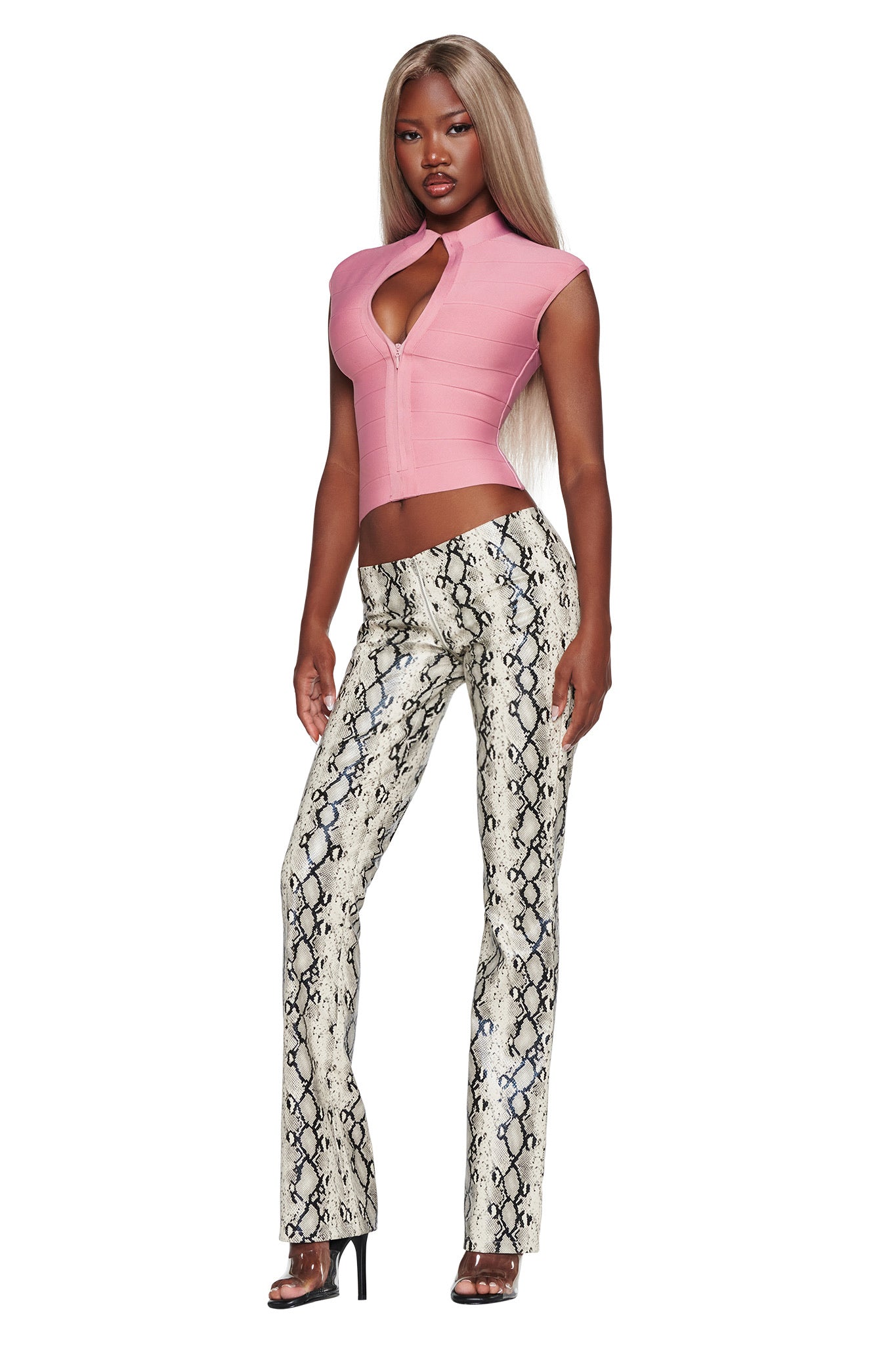 A woman with long blonde hair wears a pink, short-sleeve, keyhole crop top and the I.AM.GIA HENDRIX 2.0 PANT in Cream Snake, styled with black high heels, standing against a white background.