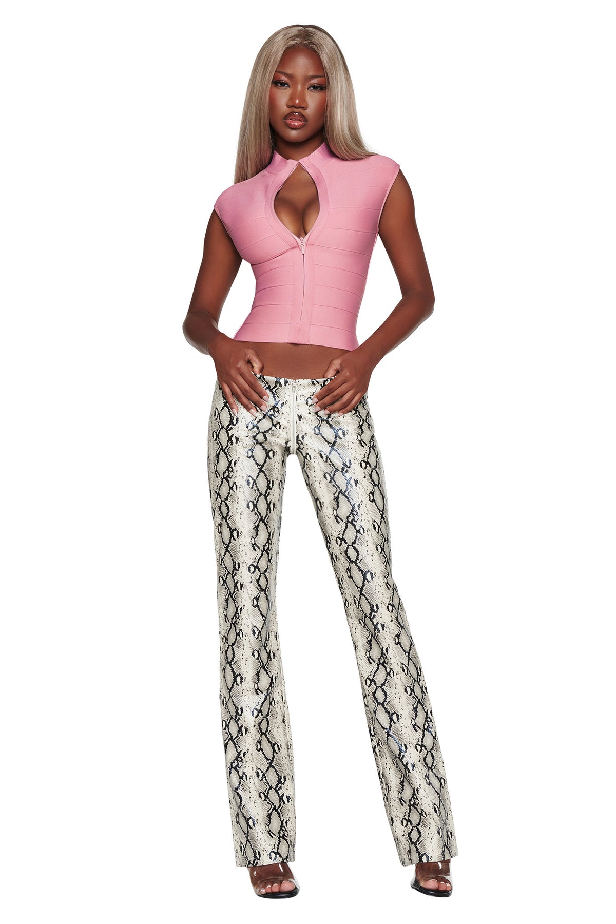 A woman stands wearing the I.AM.GIA ISELA BANDAGE TOP in dusty rose, featuring a bodycon fit and keyhole neckline, paired with high-waisted snakeskin-print pants. She has long, straight blonde hair and is wearing sandals.