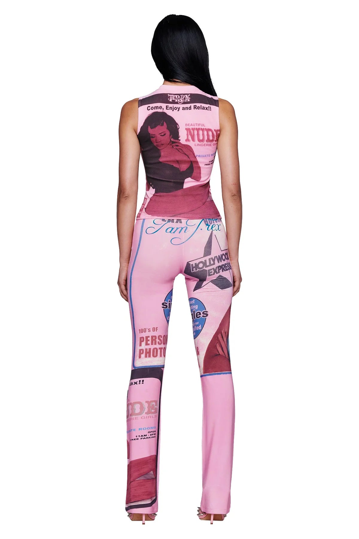 A woman stands facing away, wearing the I.AM.GIA T-REX COURTNEY TANK in pink—a sleeveless top crafted from heavyweight cotton jersey and adorned with vintage magazine covers and pin-up art in a signature I.AM.GIA placement print. She has long, straight black hair and is paired with high-heeled sandals.
