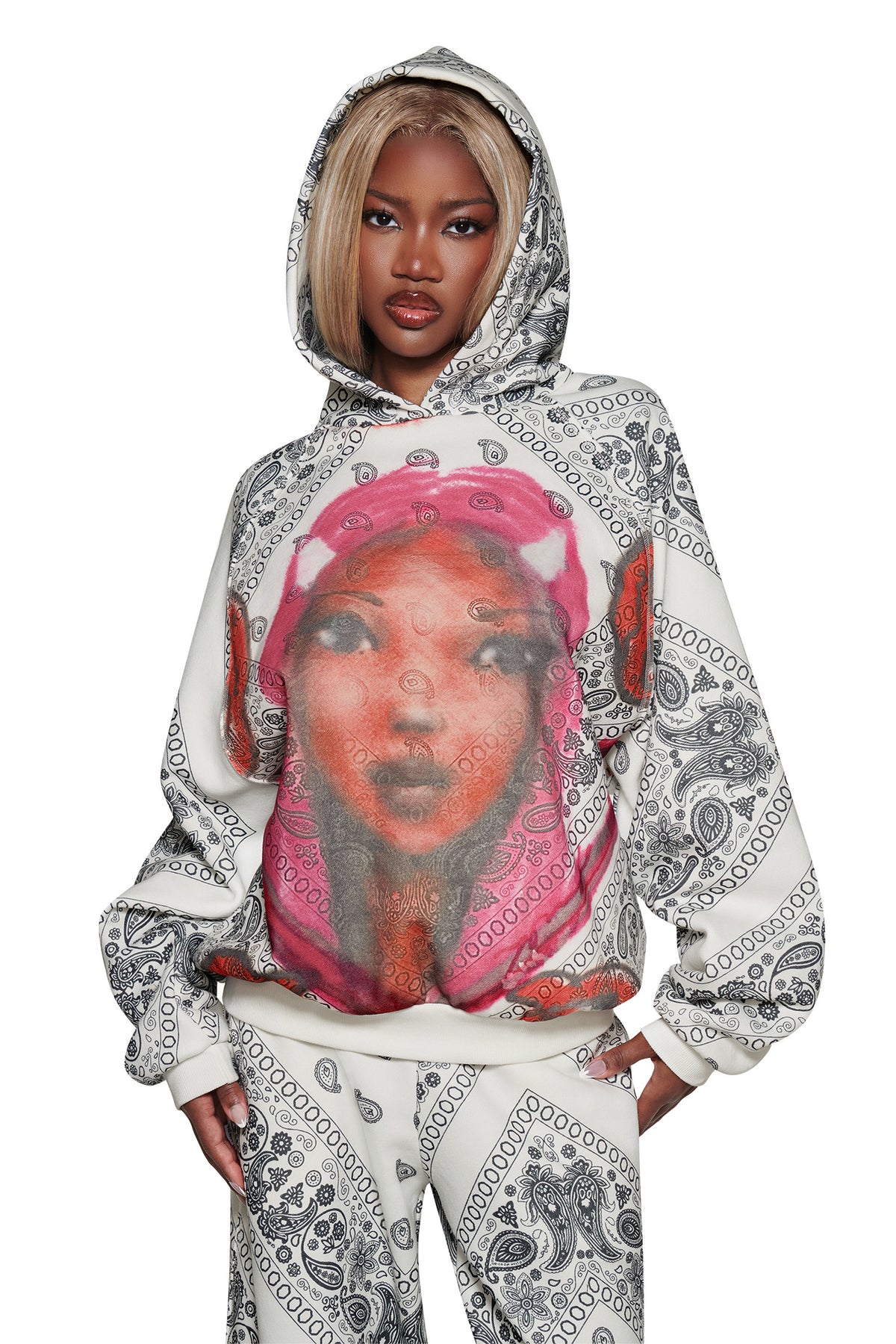 A woman with blonde hair wears the I.AM.GIA T-REX PULLOVER HOODIE - PAISLEY FACE, featuring a large colorful face print, oversized hood up, and matching pants against a white background.