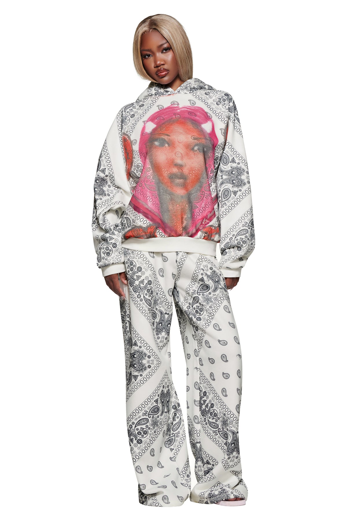 A woman with blonde hair models the I.AM.GIA T-REX PULLOVER HOODIE - PAISLEY FACE, a white fleece boxy hoodie with black paisley patterns and a large woman’s face graphic on the pink oversized hood. She stands facing forward.