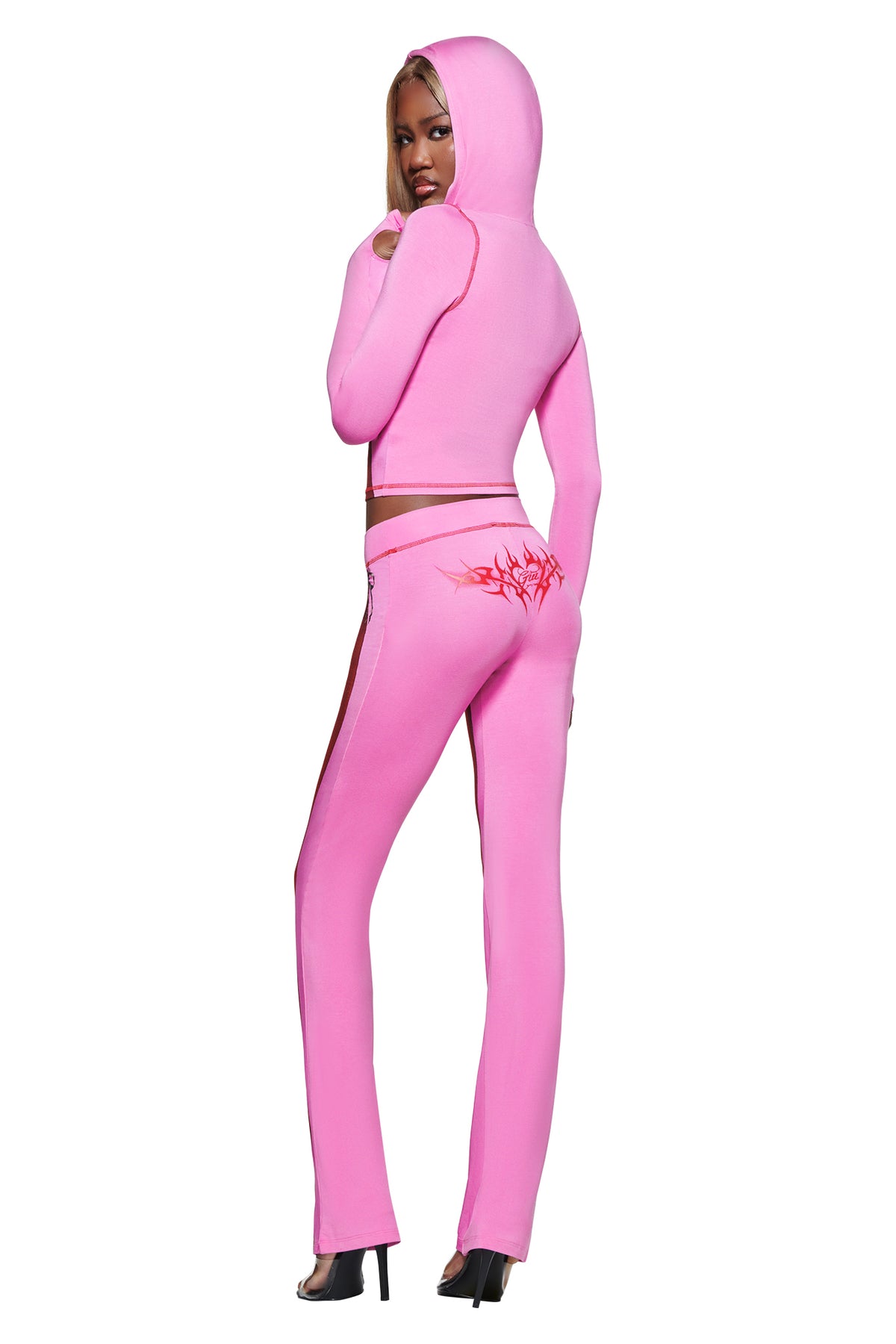 A woman stands in the T-REX SPRAY BODY BLARE HOODIE - PINK by I.AM.GIA, featuring an open back, red graphic pants, black heels, and a hint of airbrushed bikini body print for a bold look.