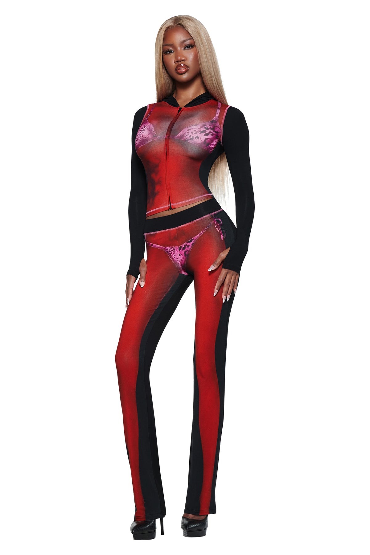 A woman with long blonde hair wears a fitted black and red sheer two-piece set with T-REX SPRAY BODY BLARE TRACKPANT - BLACK by I.AM.GIA and black heels—an effortlessly chic airport look against a white background.