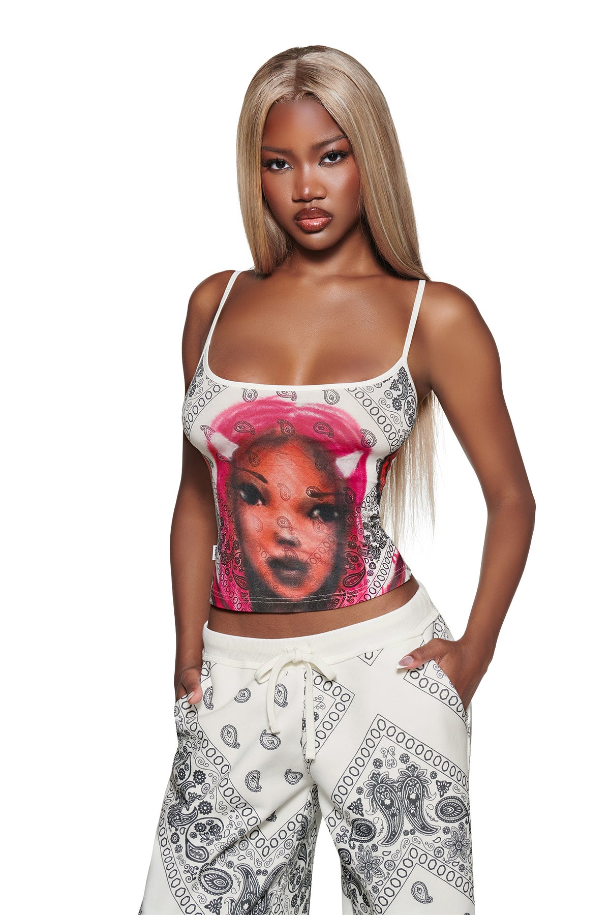 A woman with long blonde hair poses confidently in I.AM.GIA placement print bandana pants and the T-REX TANK - PAISLEY FACE. The background is plain white.