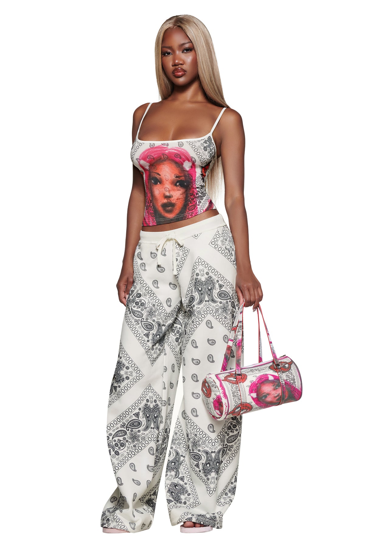 Woman with long blonde hair wears the I.AM.GIA T-REX TANK - PAISLEY FACE, white patterned wide-leg pants, and holds a pink handbag while standing against a white background.