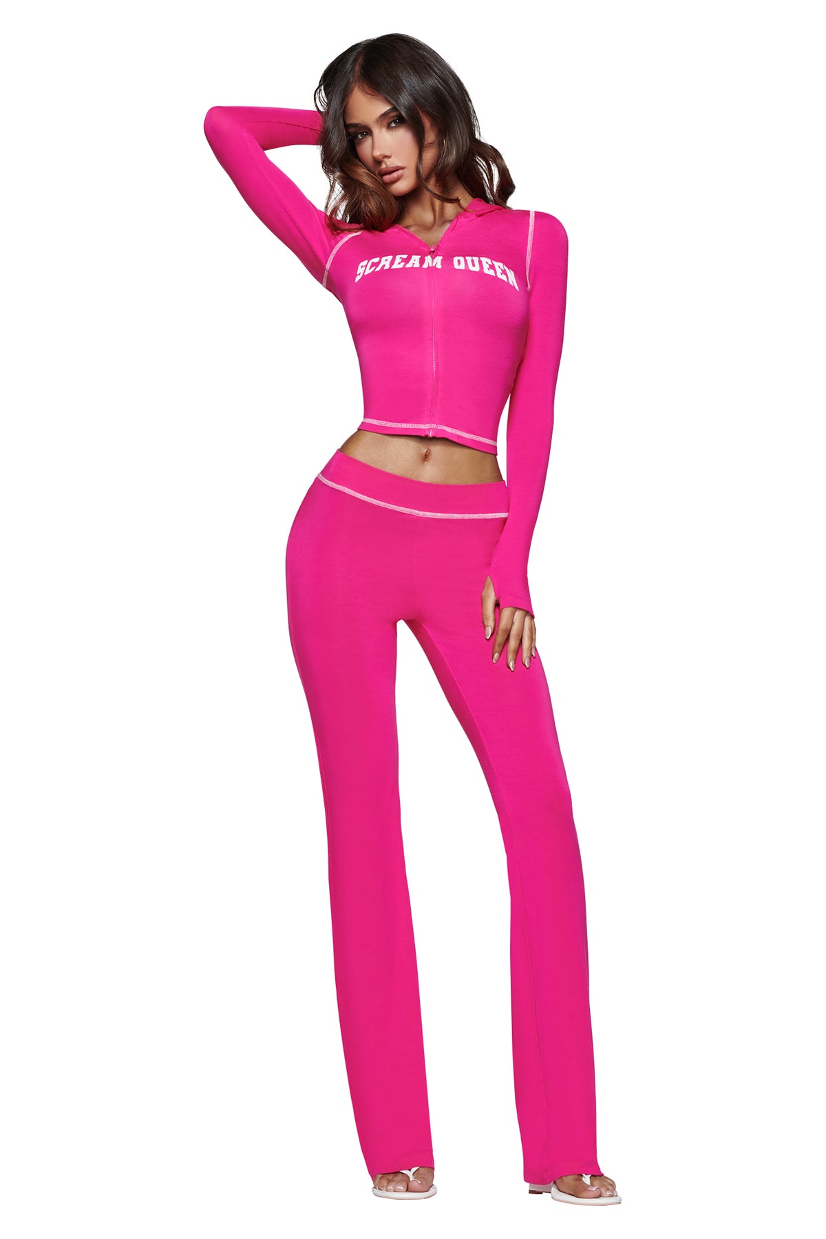 A woman with long hair, wearing the I.AM.GIA BLARE HOODIE in hot pink featuring a bold “Scream Queen” graphic, stands with one arm raised to her head and the other at her side. She completes the look with clear-heeled shoes.