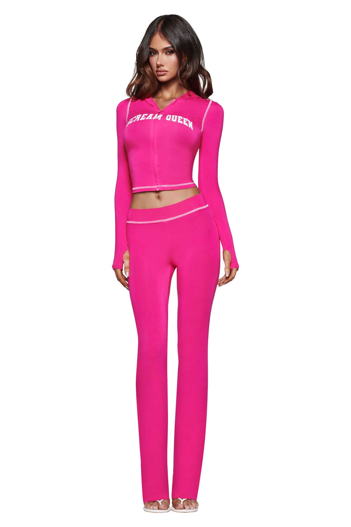 A woman stands against a white background wearing the I.AM.GIA BLARE TRACKPANT - HOT PINK SCREAM QUEEN, styled with a matching long-sleeve crop top featuring a bold Scream Queen graphic and clear high-heeled shoes.