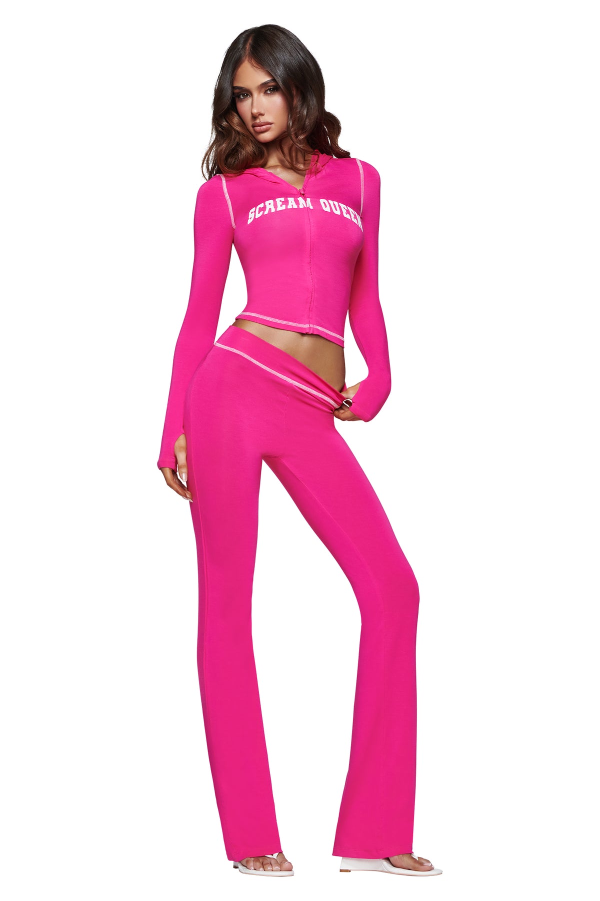A woman models the I.AM.GIA BLARE HOODIE - HOT PINK SCREAM QUEEN, a vibrant pink long-sleeve piece emblazoned with the bold “Scream Queen” graphic, paired with matching flared pants. She poses confidently with one hand on her hip and the other adjusting her waistband against a white background.