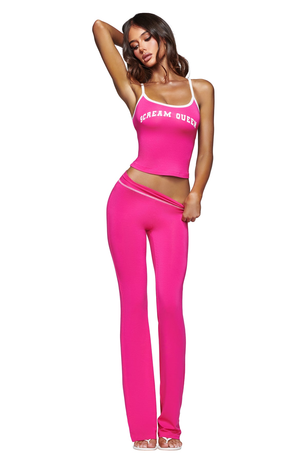 A woman poses barefoot against a white background wearing the I.AM.GIA BLARE TRACKPANT - HOT PINK SCREAM QUEEN set, which features a hot pink spaghetti strap crop top emblazoned with a bold “Scream Queen” graphic and matching high-waisted, flared track pants.