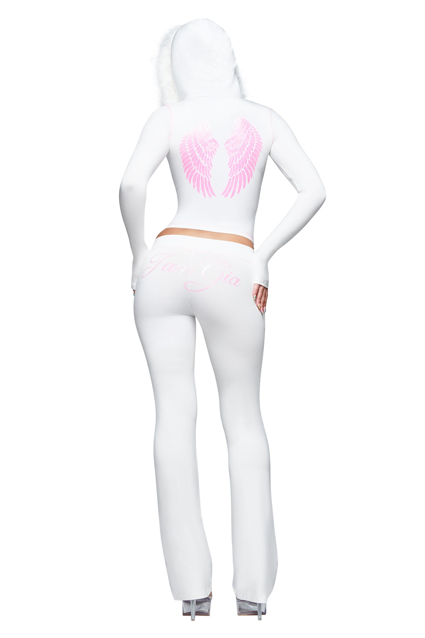 Dressed in the I.AM.GIA BLARE TRACKPANT - WHITE : ANGEL, featuring pink angel wings printed on the back and script detailing, this individual stands on clear platform heels with one hand on their hip. The low-rise flared trackpants and signature design elements embody the iconic I.AM.GIA ANGEL aesthetic.