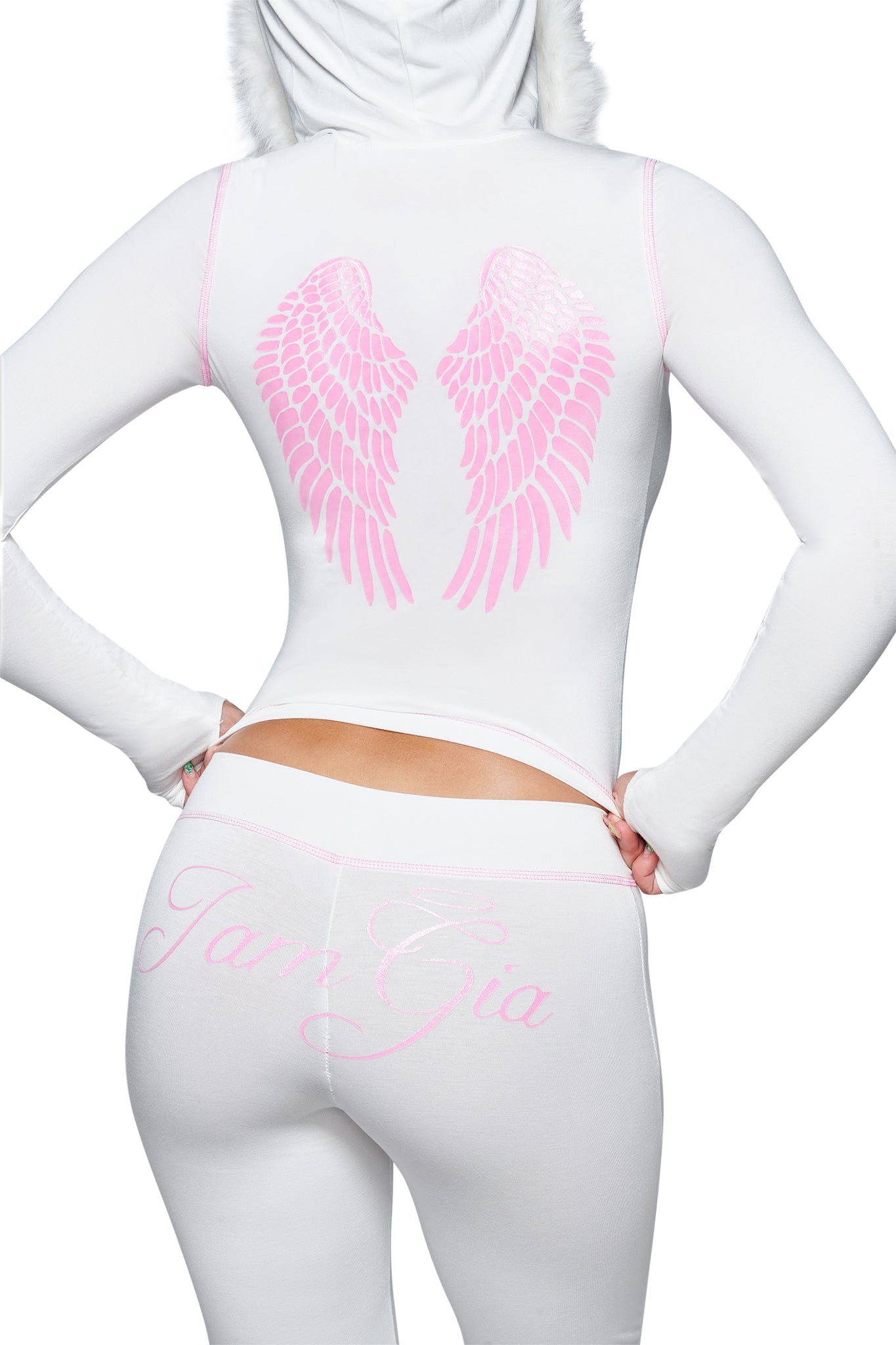 A person is shown from behind against a white background, wearing the I.AM.GIA BLARE TRACKPANT - WHITE : ANGEL, a white hooded outfit with pink angel wings and cursive “IamGia” text printed on the back of the pants. Featuring the signature ANGEL look from I.AM.GIA, these low-rise flared pants add a bold touch to the striking ensemble.