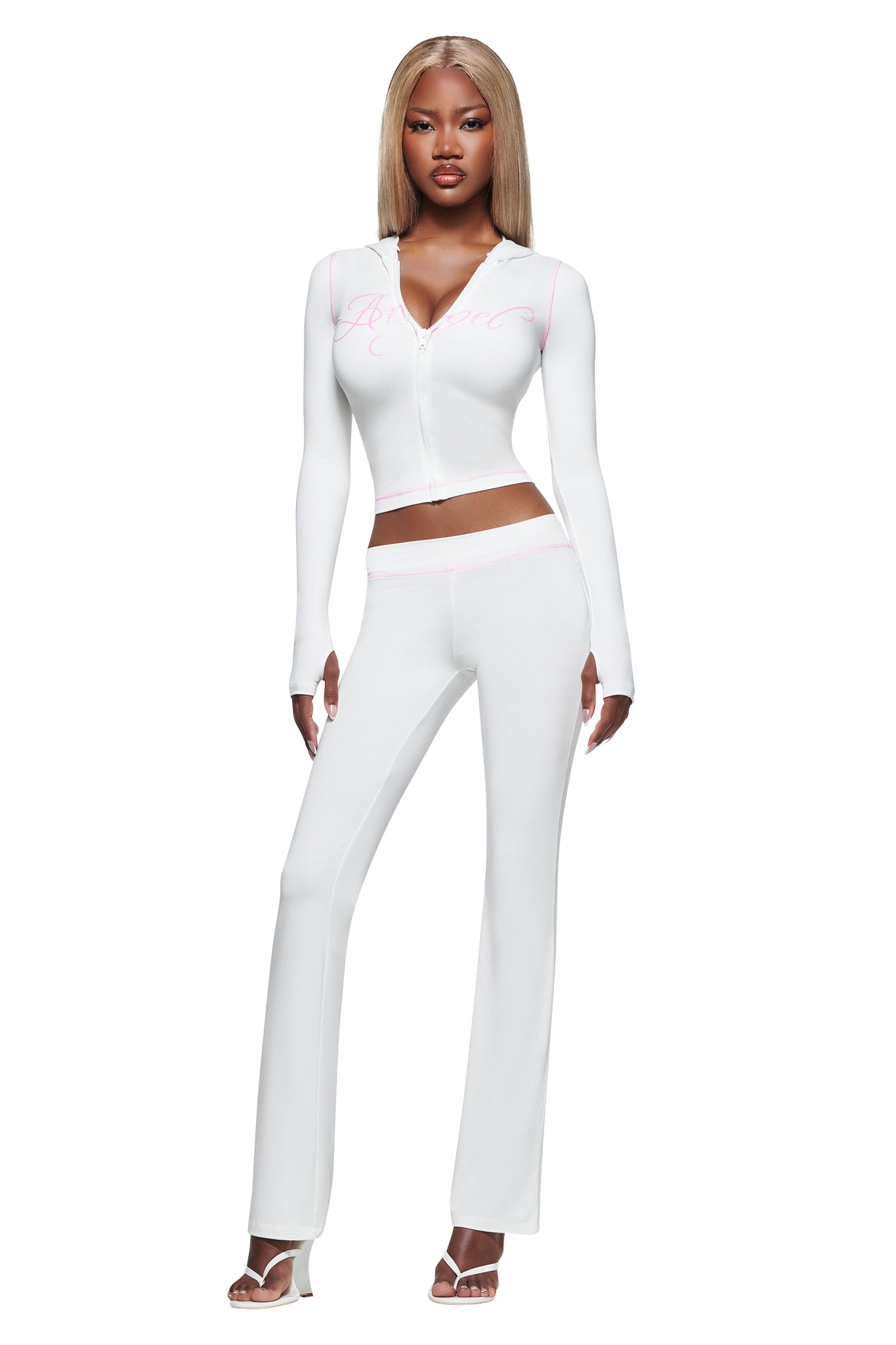 A woman with long blonde hair wears a fitted white zip-up top with pink script, paired with I.AM.GIA’s BLARE TRACKPANT - WHITE : ANGEL low rise flared pants. She stands confidently in white heeled sandals against a plain white background.