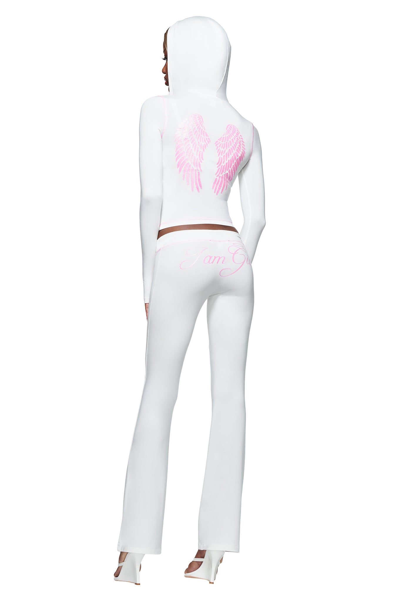 A person wearing the I.AM.GIA BLARE FUR TRIM HOODIE in white, featuring pink angel wings on the back, paired with pants that have “I am G” written on them and white high-heeled shoes, stands with their back to the camera.