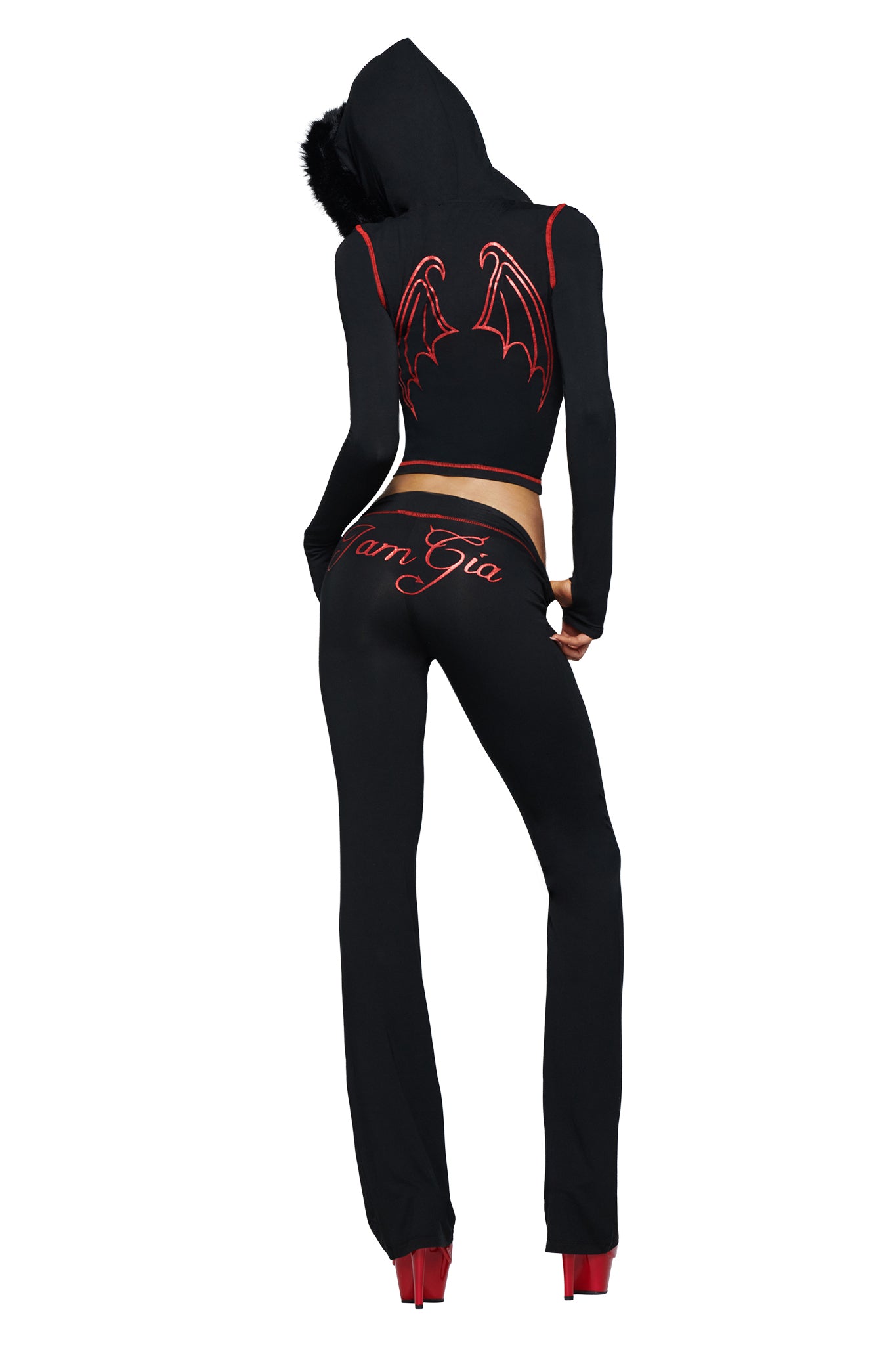 Model stands facing away from the camera, dressed in a black hooded outfit featuring red bat wings and the devil GIA logo on the back. She wears I.AM.GIA’s BLARE TRACKPANT - JET BLACK : DEVIL, emblazoned with “I AM GIA” lettering down the leg, paired with bold red platform heels.