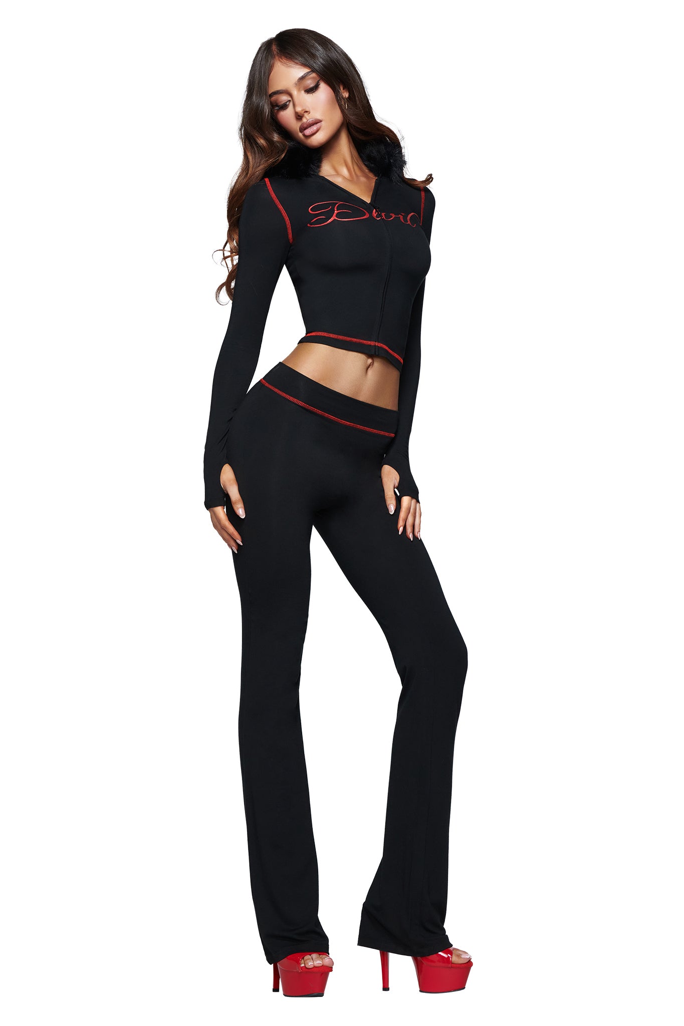 A woman poses in a black two-piece outfit with striking red trim and high platform heels. The long-sleeved, cropped top showcases the iconic devil GIA logo in bold red lettering across the chest. Her look is completed with the I.AM.GIA BLARE TRACKPANT - JET BLACK : DEVIL, a high-waisted, fitted pant that blends sleek style with an athletic edge.