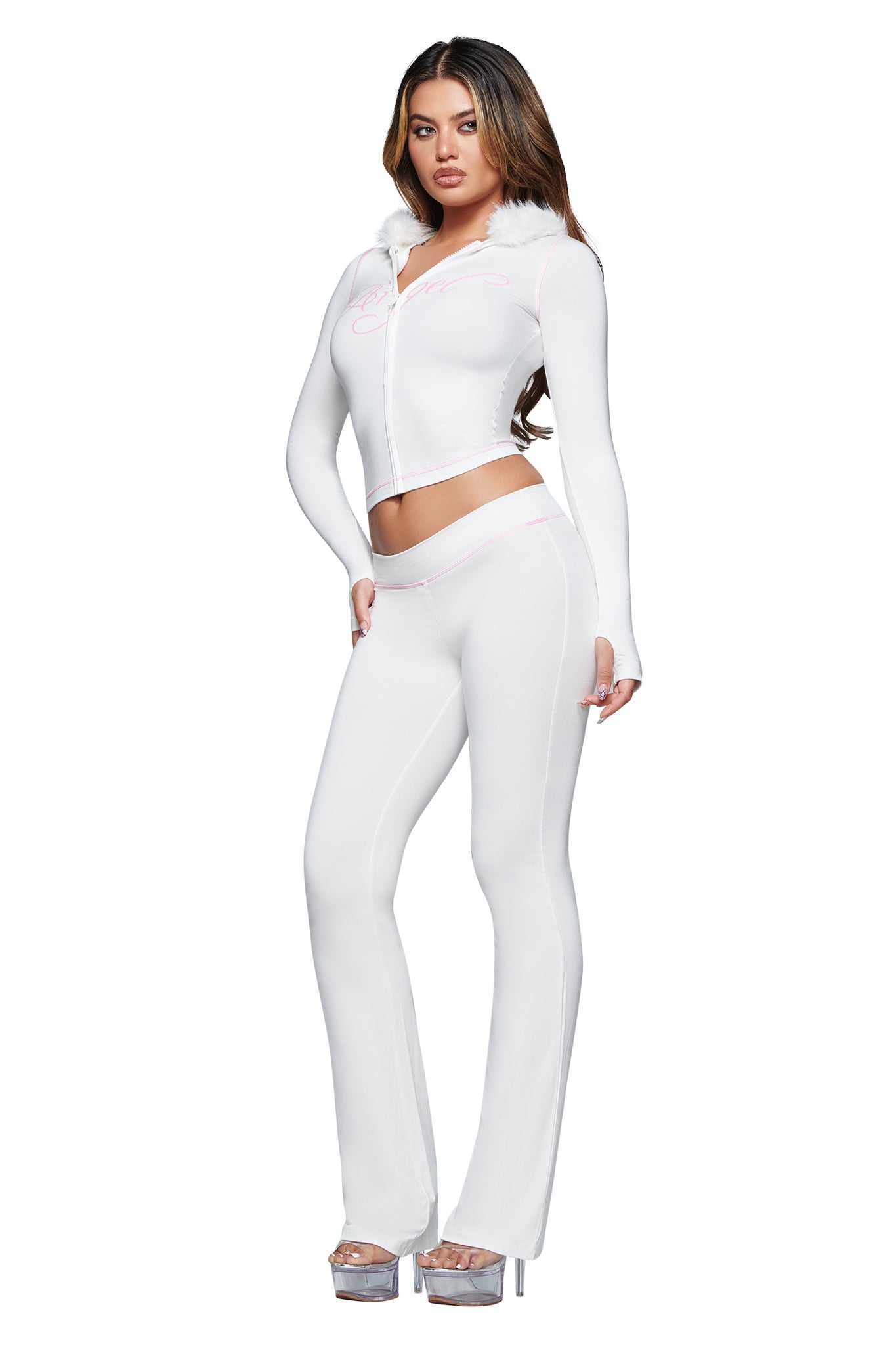 A woman poses in the I.AM.GIA BLARE TRACKPANT - WHITE : ANGEL, paired with a fitted white zip-up hoodie with fur trim and silver high-heeled platform shoes, standing against a white background.