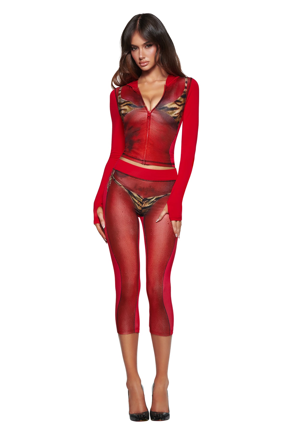 A woman in a red fitted zip-up top and I.AM.GIA T-REX SPRAY BODY BLARE CAPRI - RED with sheer panels, metallic gold pattern, and black heels poses against a white background.