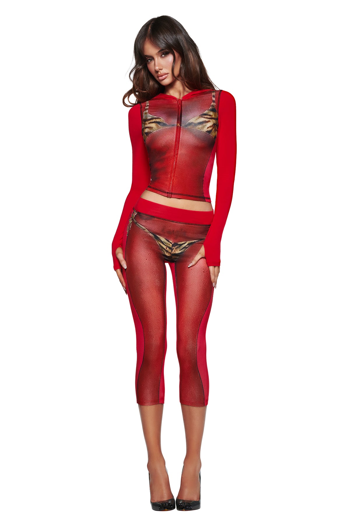 A woman in a red fitted outfit with sheer panels, gold accents, and T-REX SPRAY BODY BLARE CAPRI - RED by I.AM.GIA stands facing forward in black heels against a white background.