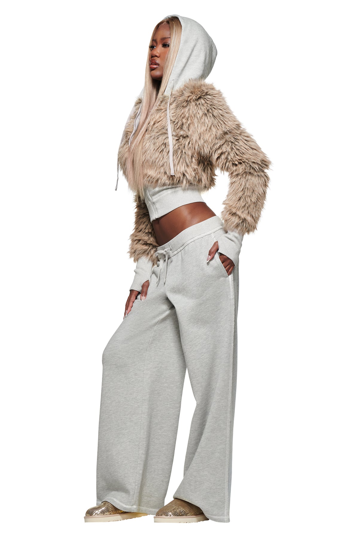 Wearing the I.AM.GIA KOKO TRACKPANT in Heather Grey styled in a wide leg silhouette, she stands confidently with hands on hips, pairing the look with a furry tan cropped hoodie and gold sneakers, gazing off to the side.