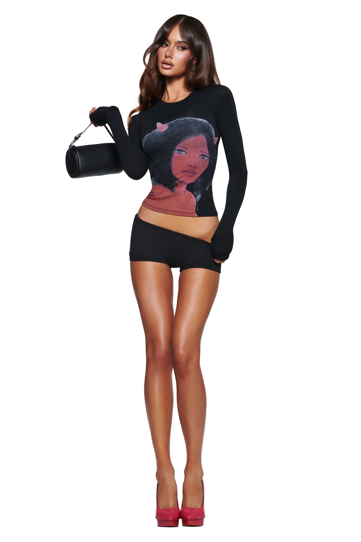 A woman in a black long-sleeve top with an anime face print, T-REX BLARE 2.0 SHORT - BLACK by I.AM.GIA, pink heels, and a black clutch poses against a white background.