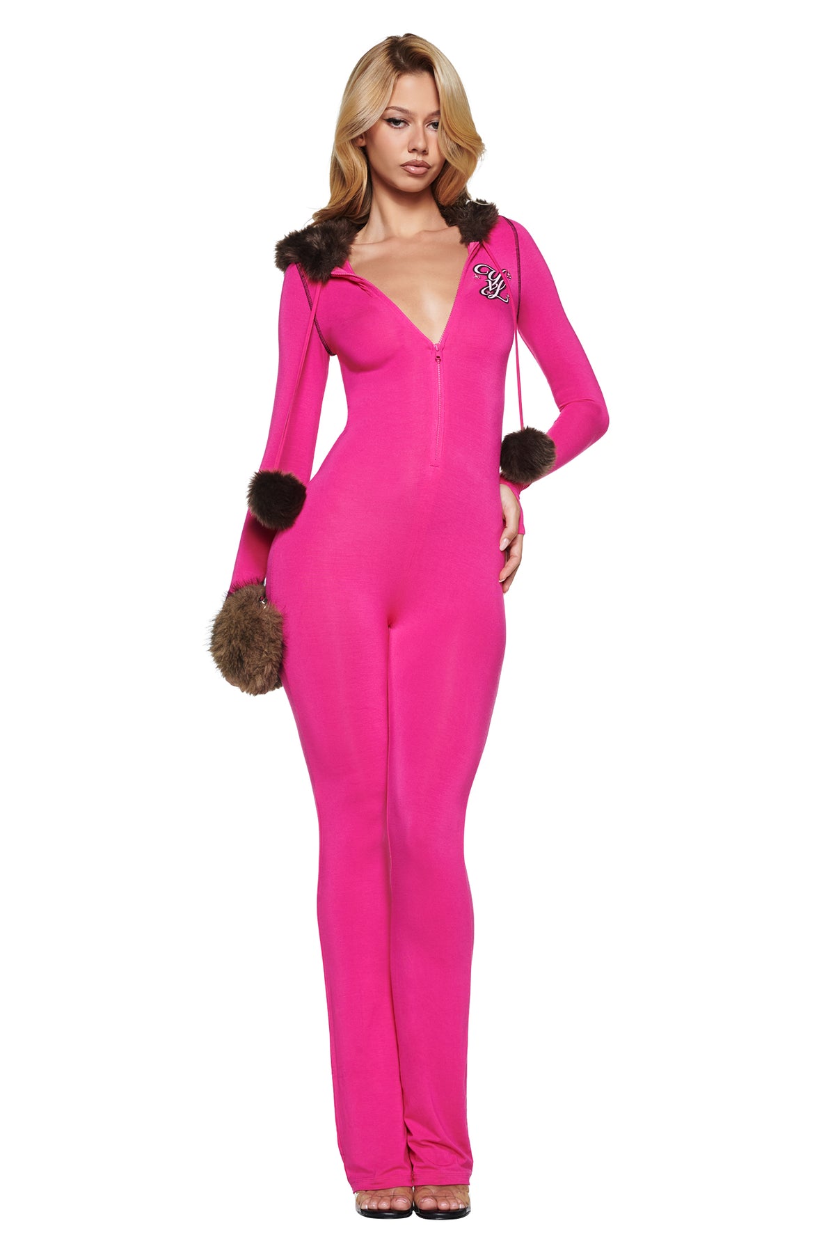 A person wearing a bright pink YVL BLARE FUR LINED JUMPSUIT with a deep V-neck and flare leg pants, featuring faux fur trim on the hood and cuffs, and small fur pom-poms on the drawstring.