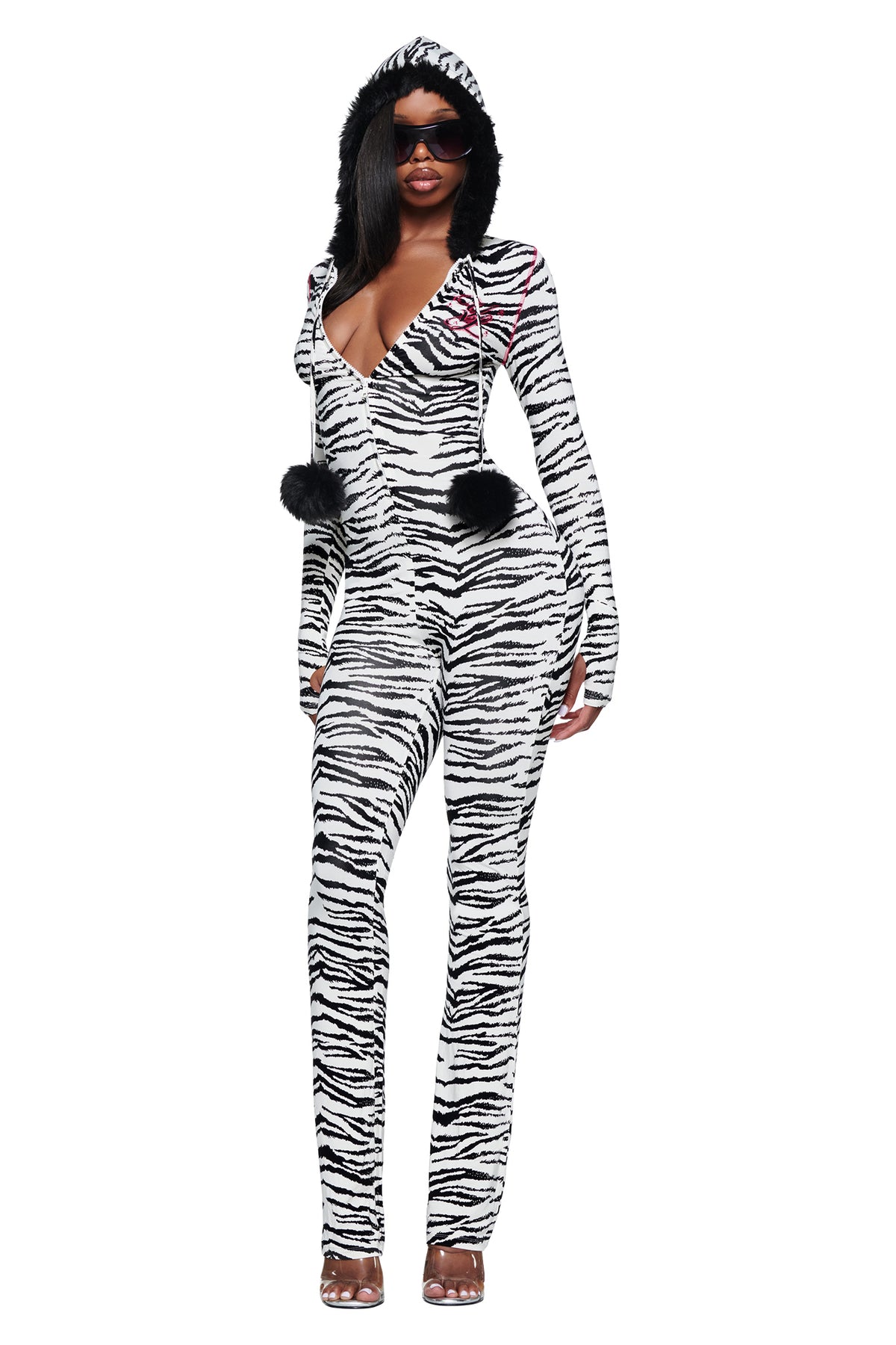 A model wears the YVL BLARE FUR LINED JUMPSUIT - ZEBRA in a zebra print, featuring a black faux fur-lined hood with two black pom-poms hanging from the drawstrings.