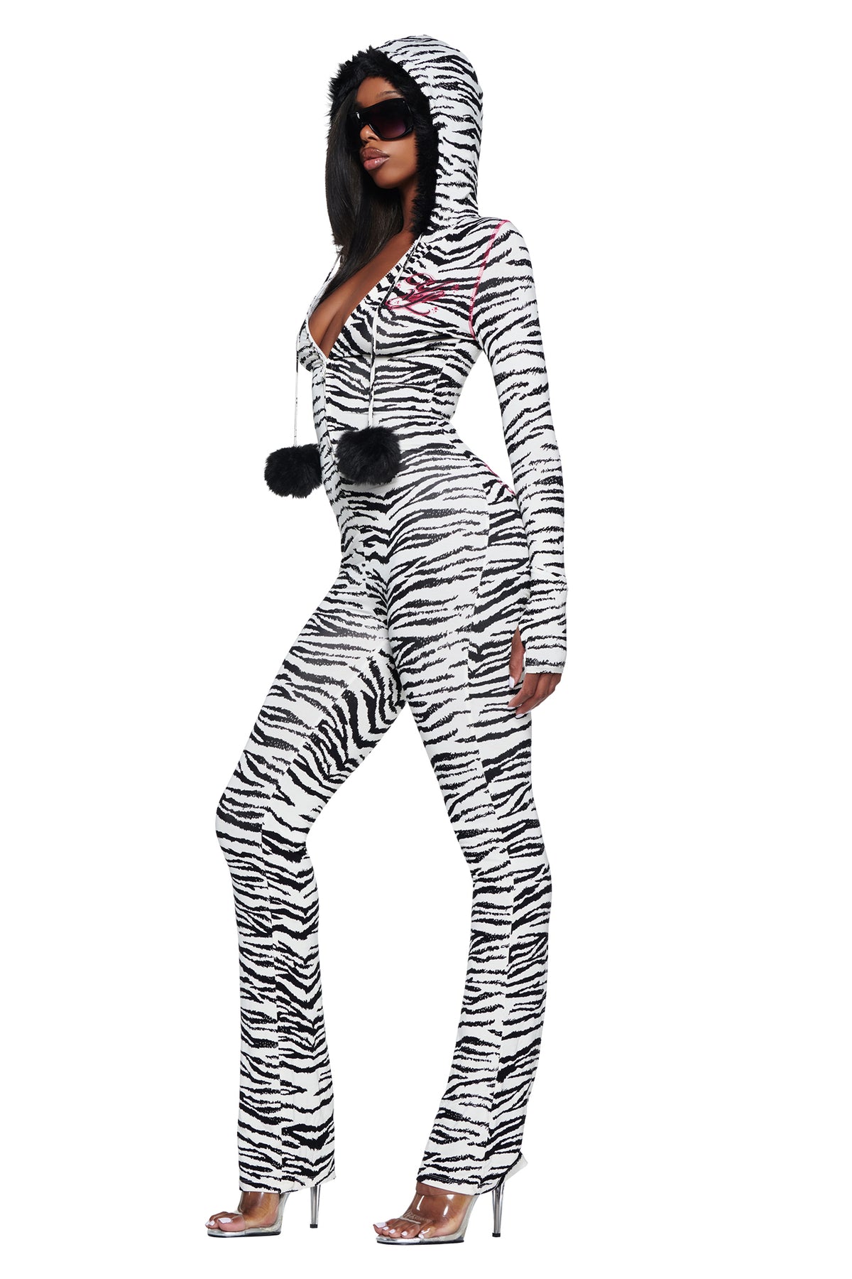 A person wearing an I.AM.GIA YVL BLARE FUR LINED JUMPSUIT - ZEBRA, featuring a long-sleeved zebra print design with a faux fur hood and black pom-poms hanging from the front zipper.