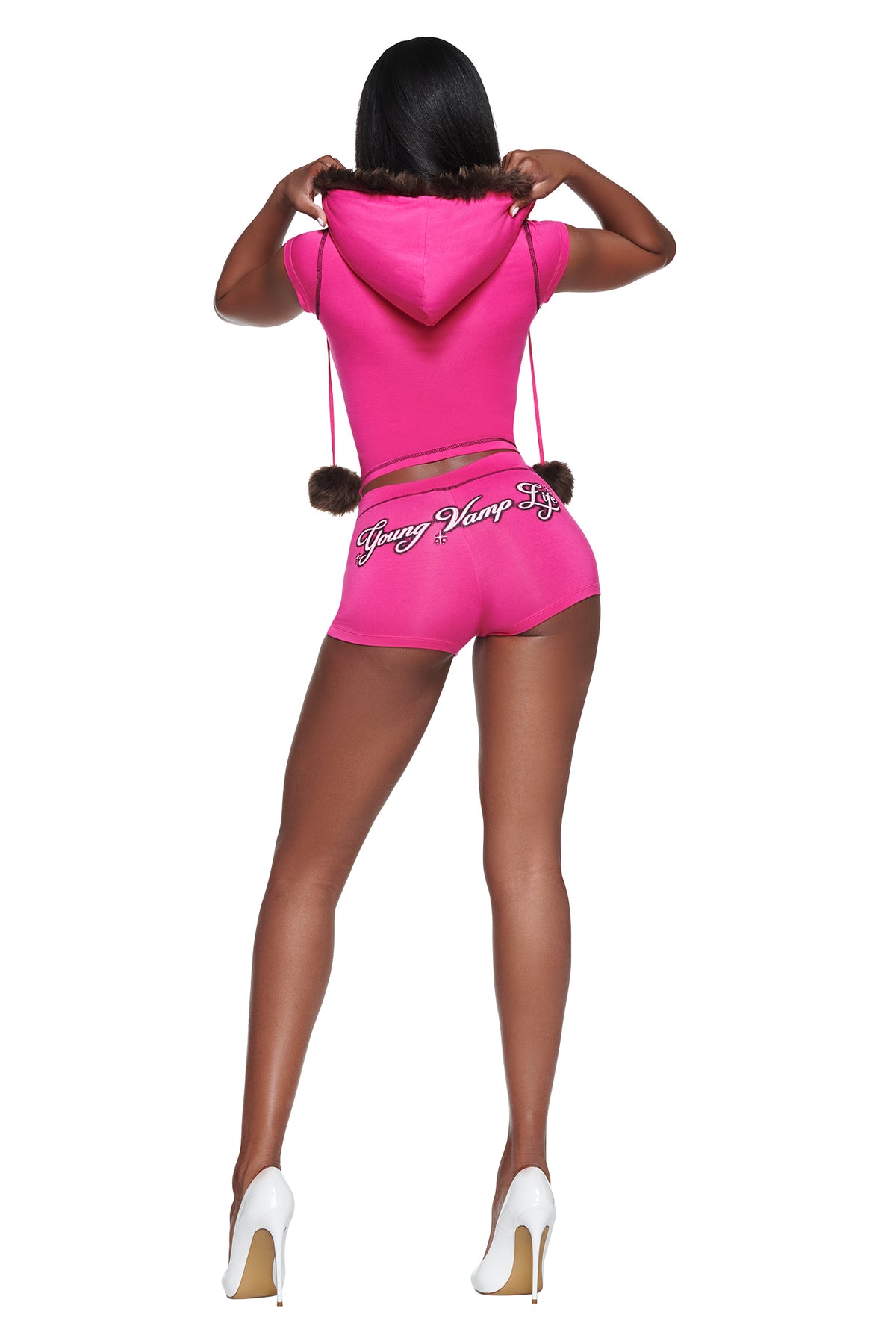 A woman seen from behind wearing a pink I.AM.GIA YVL BLARE FUR LINED CAP SLEEVE HOODIE with matching short shorts featuring “Young Vamp Life” embroidery, paired with white high heels.