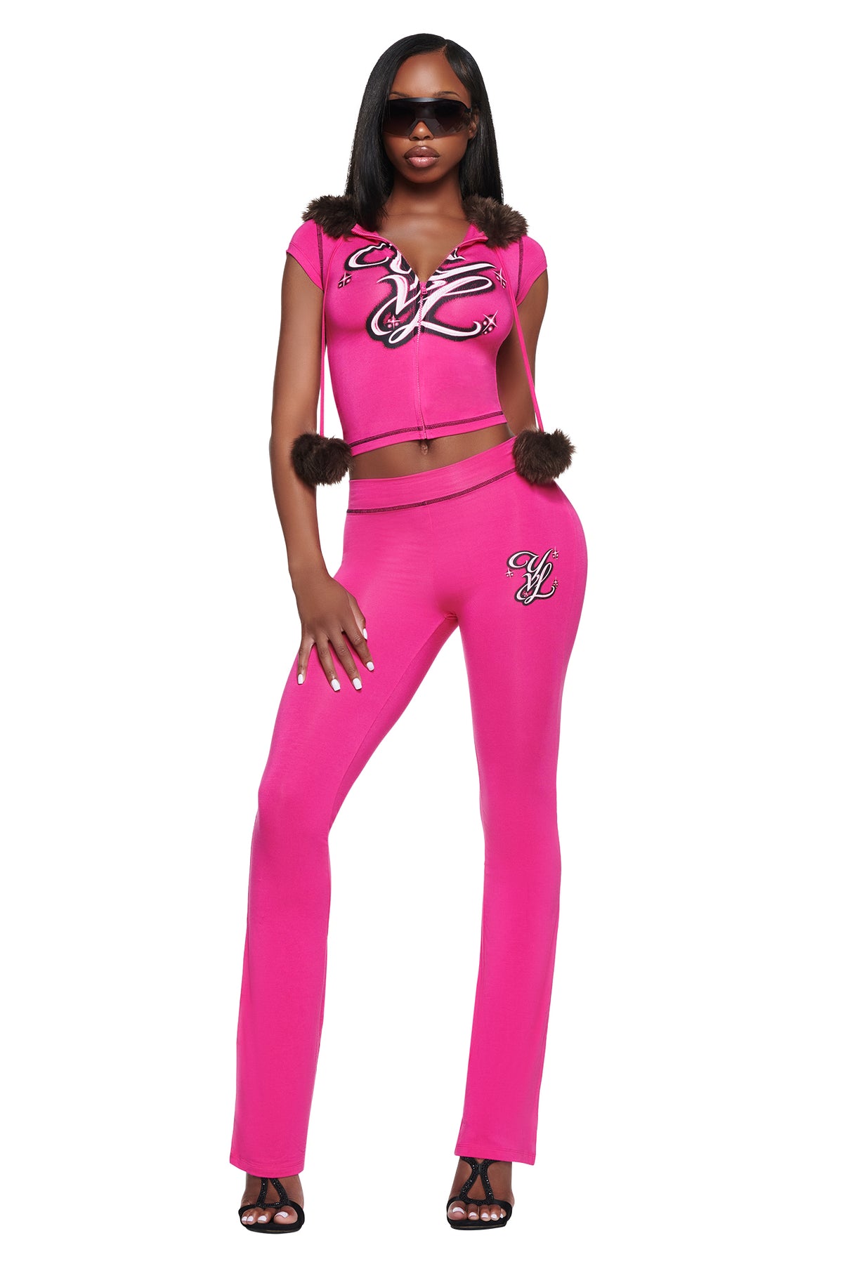 A model wears a matching bright pink cropped hoodie with a brown fur-trimmed hood and slim-fit YVL BLARE TRACKPANT - PINK, both featuring embroidered logo details.