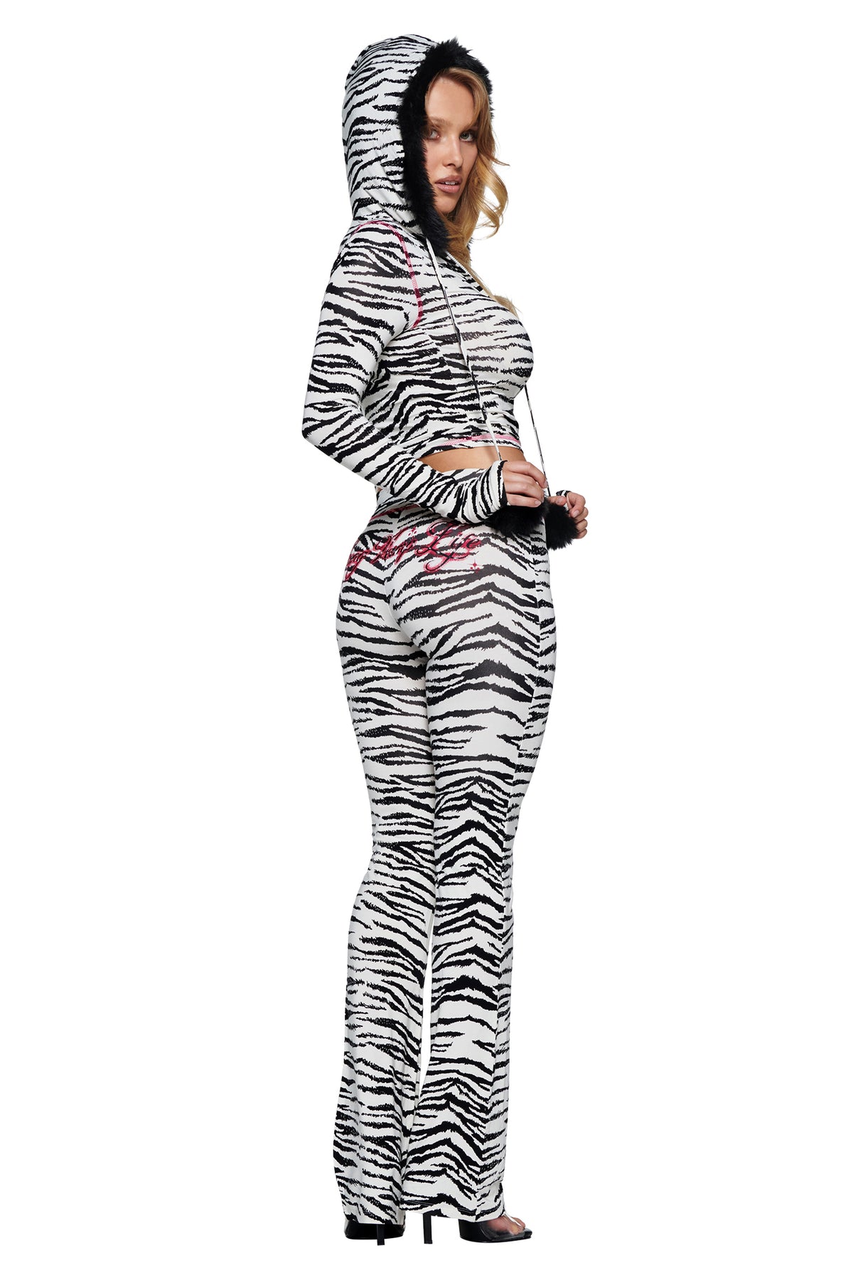 A person wearing a zebra-print hooded long-sleeve crop top and matching YVL BLARE TRACKPANT - ZEBRA flared pants with pink embroidered lettering on the back.