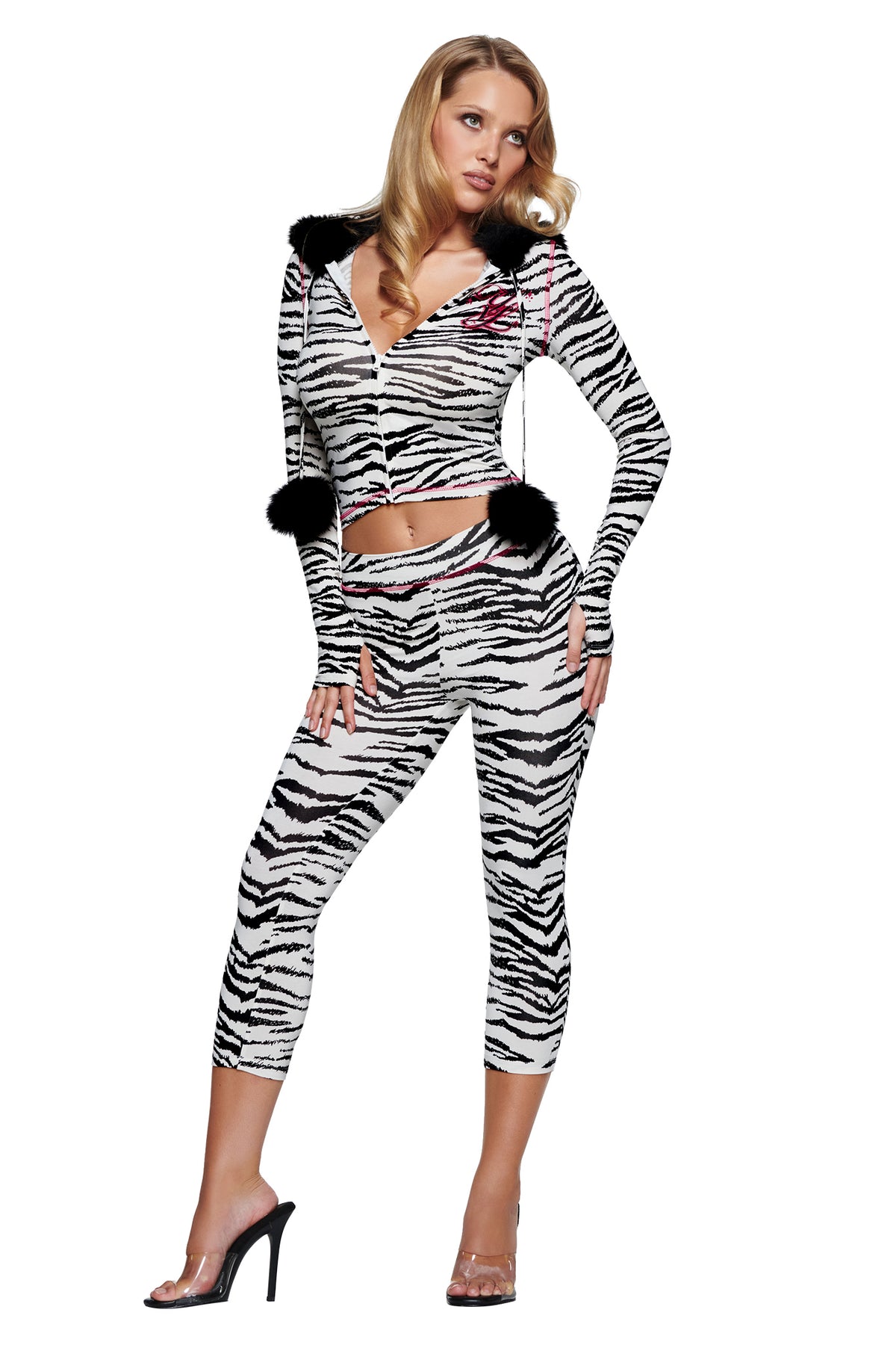 A woman wears a zebra-print hooded zip-up crop top with black pom-pom drawstrings and matching YVL BLARE CAPRI - ZEBRA leggings, paired with clear high-heeled sandals.