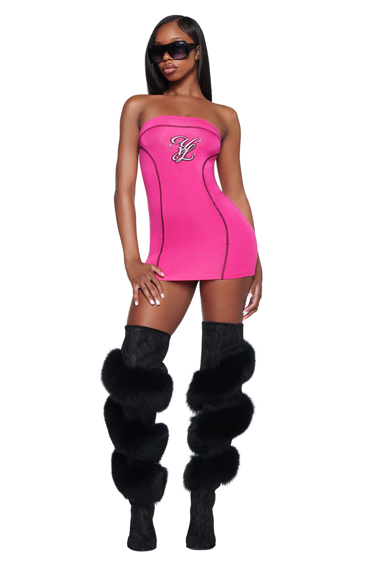 A model wears a hot pink I.AM.GIA YVL BLARE TUBE MINI DRESS with a dark logo on the chest and black knee-high boots with fur trim.
