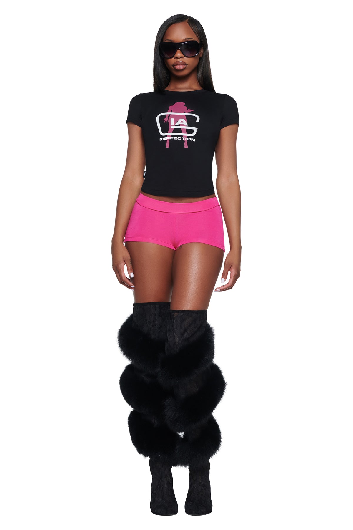 A woman wearing a black YVL T-SHIRT - BLACK GIA, bright pink shorts, and knee-high boots featuring black faux fur trim.