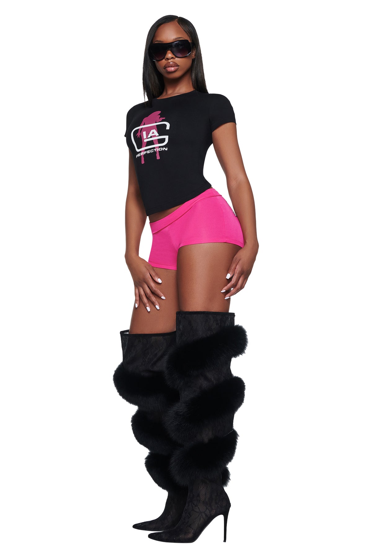 A person wearing a black I.AM.GIA YVL T-SHIRT - BLACK GIA graphic t-shirt, bright pink shorts, and black fur-trimmed, knee-high boots.