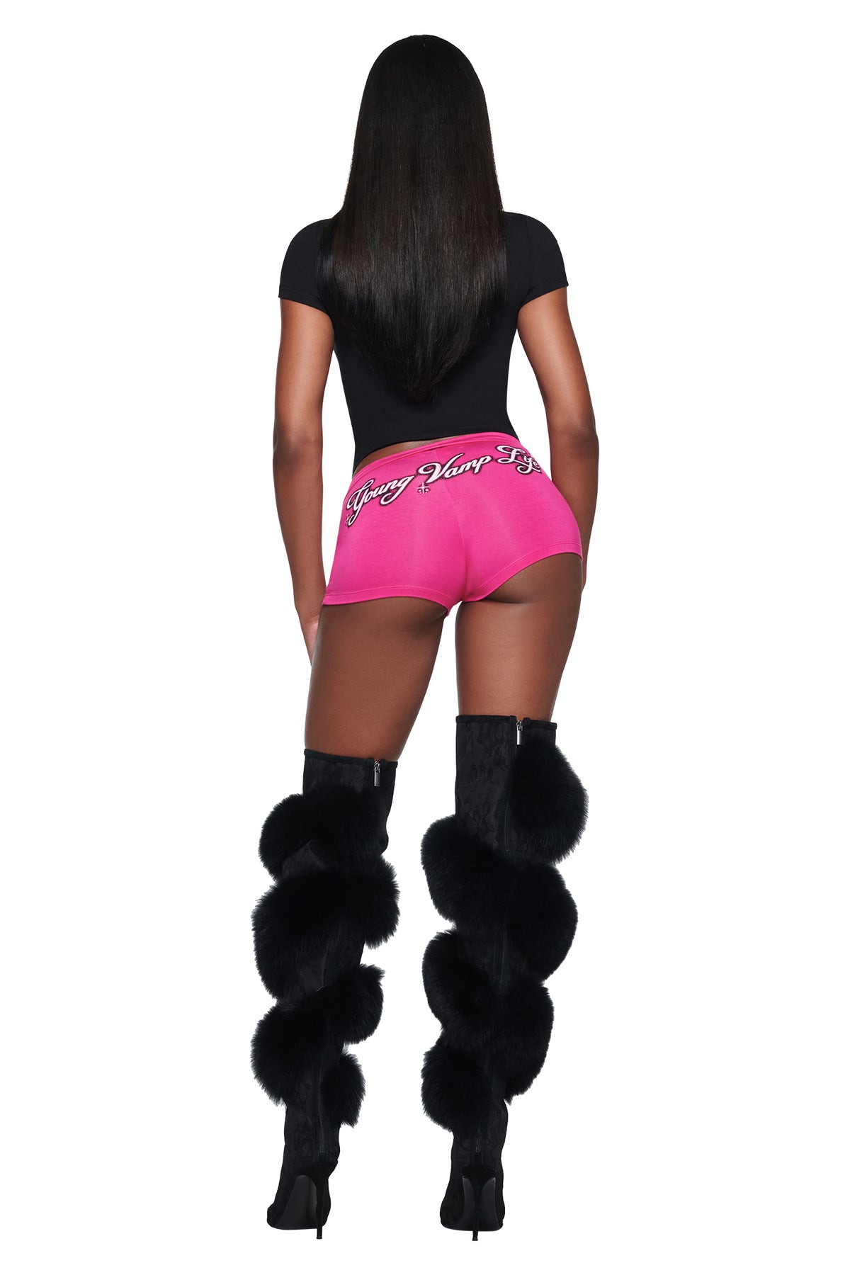 A person seen from behind wears a black short-sleeved YVL T-SHIRT - BLACK GIA, pink shorts with “Young Vamp” embroidered on the back, and black knee-high boots decorated with large fur pom-poms.
