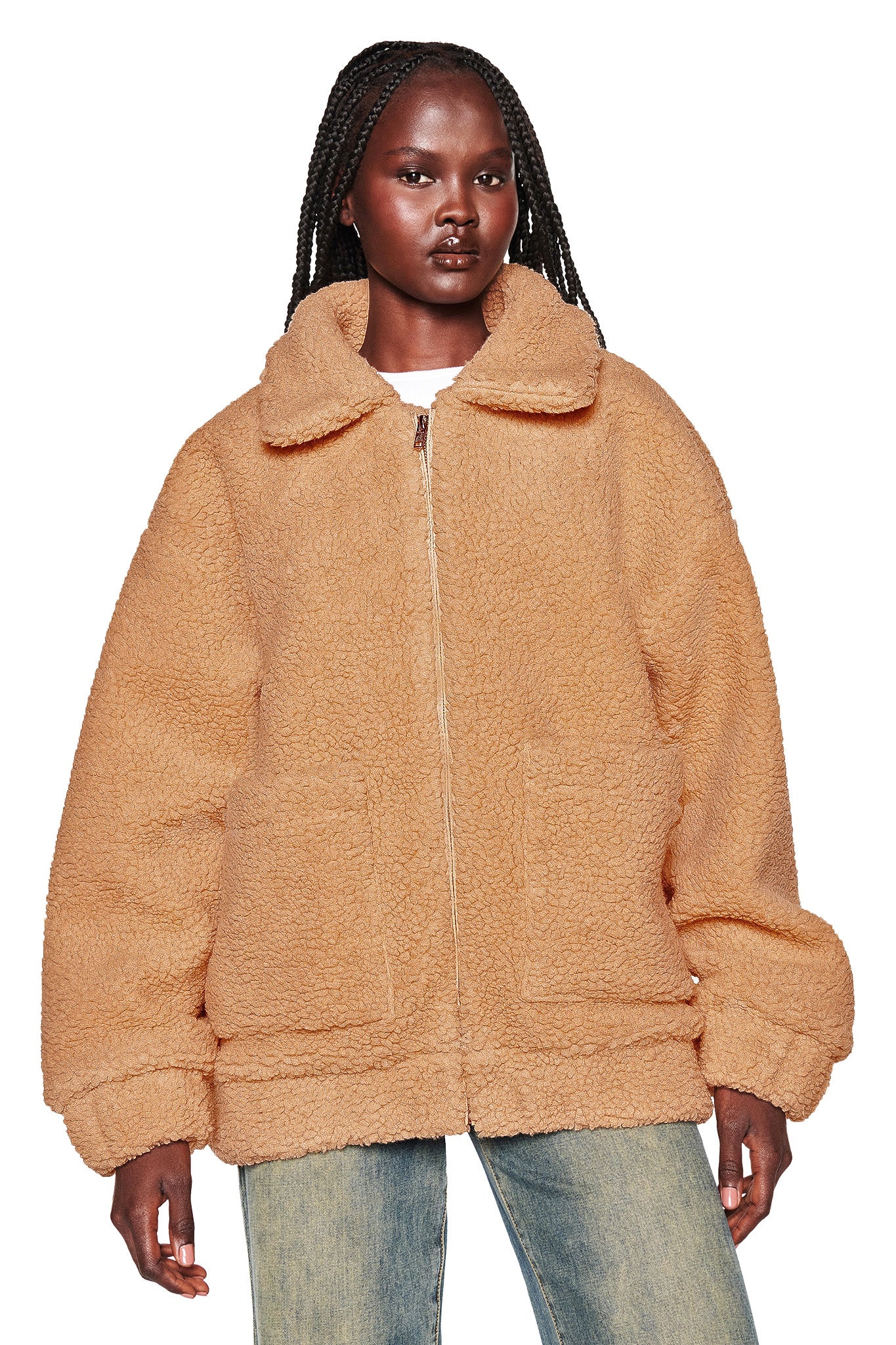 A person with long, braided hair is wearing an oversized caramel PIXIE COAT from I.AM.GIA, featuring a high collar and front pockets. Paired with light blue jeans, their expression remains neutral against the plain white background.