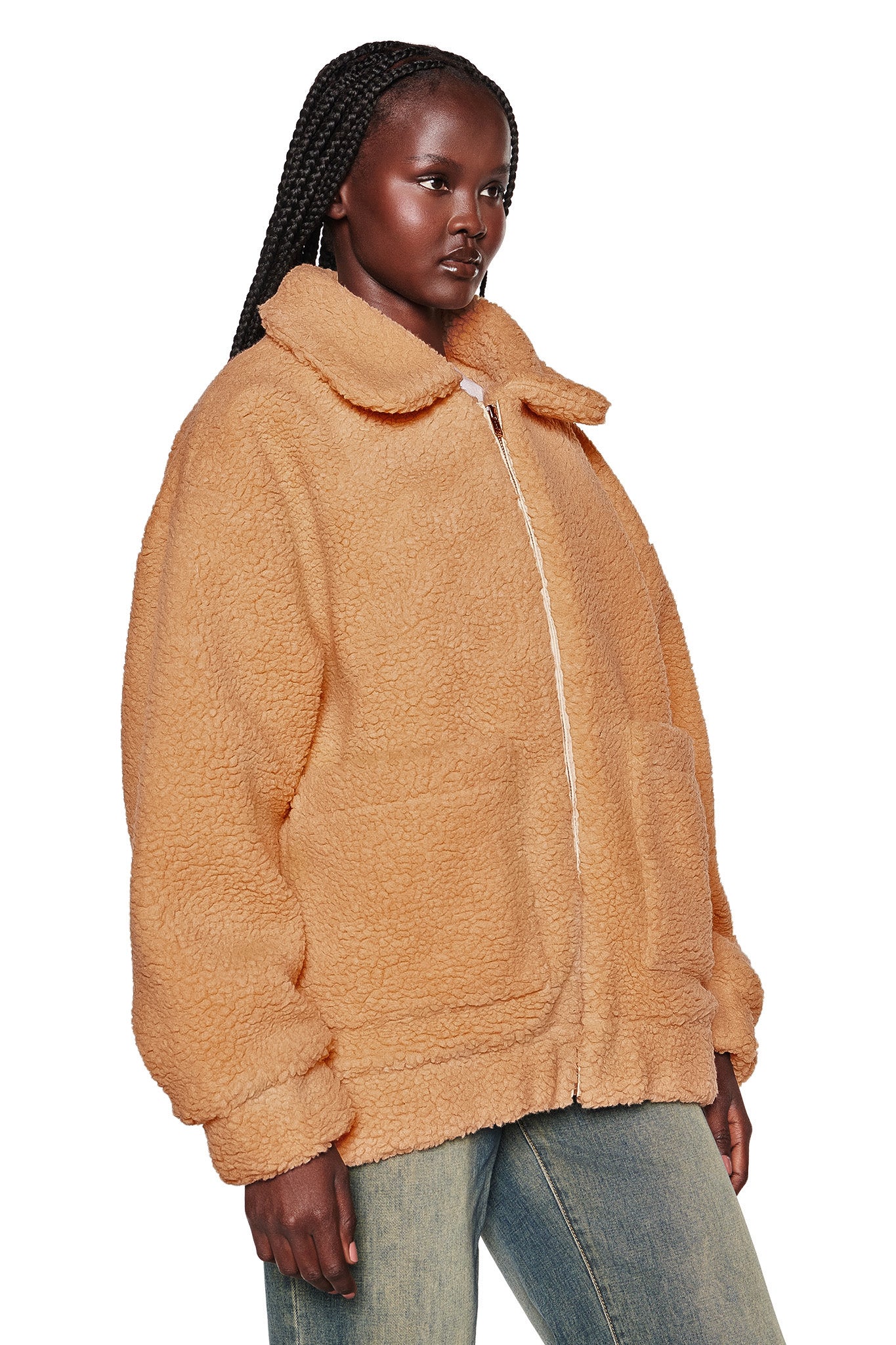 A person with long braided hair is wearing a light brown, oversized I.AM.GIA PIXIE COAT in caramel, featuring large front pockets. The zippered coat has a wide collar. They are also wearing blue jeans and standing against a plain white background.